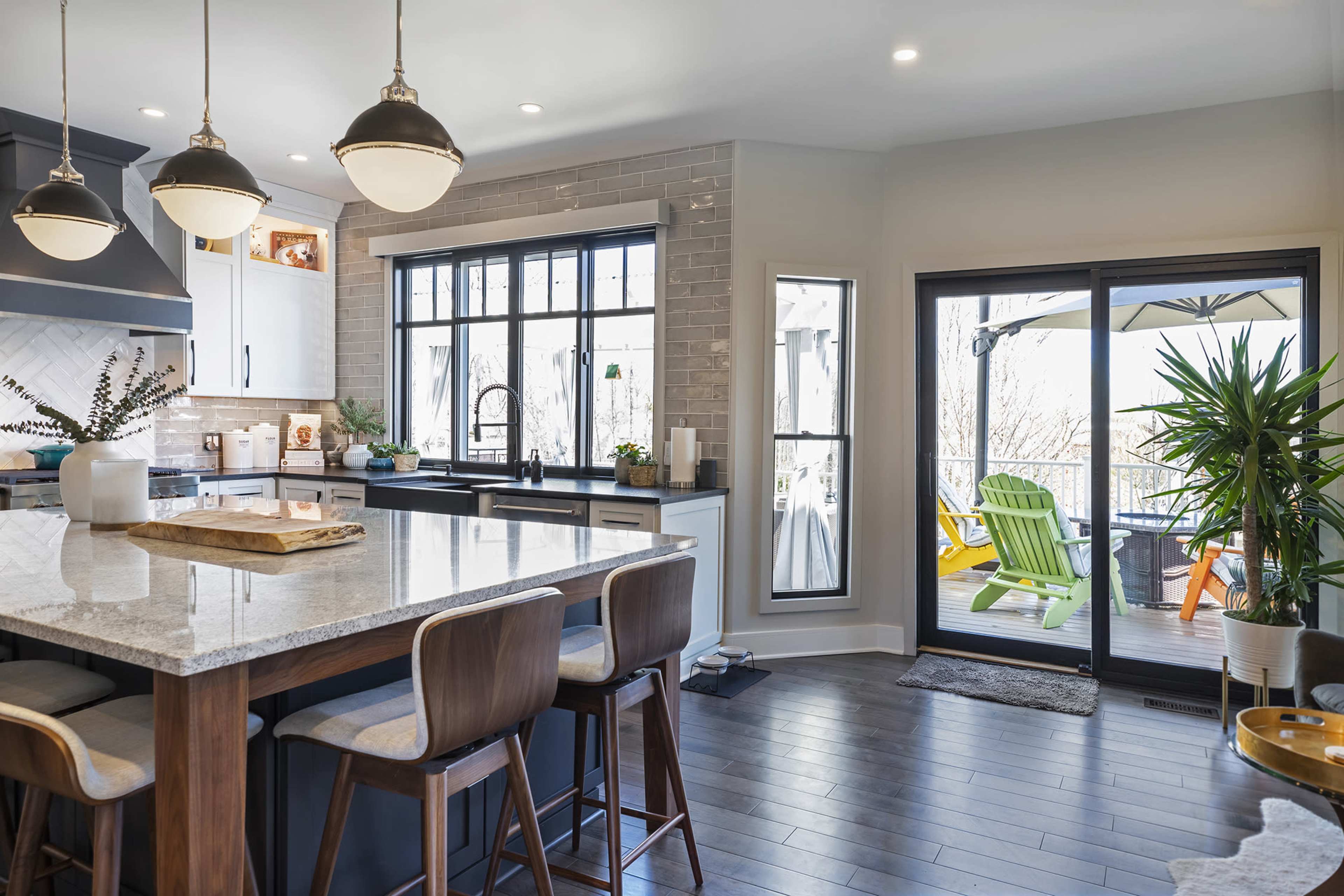 A modern kitchen features a large island with wooden stools, gray cabinets, and large windows overlooking a deck with colorful chairs.