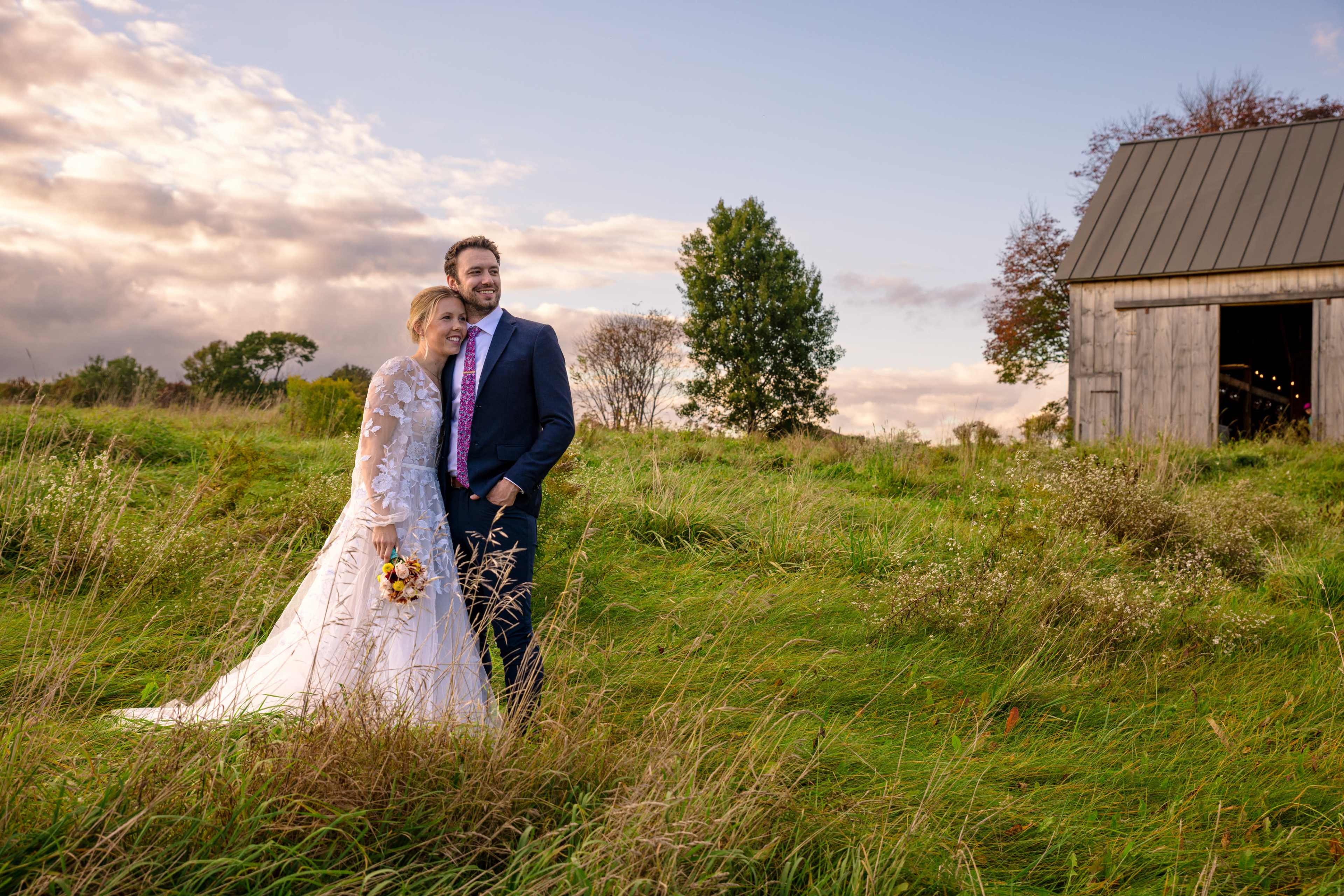 A bride in a white wedding dress and a groom in a suit stand together in a grassy field near a barn under a cloudy sky.