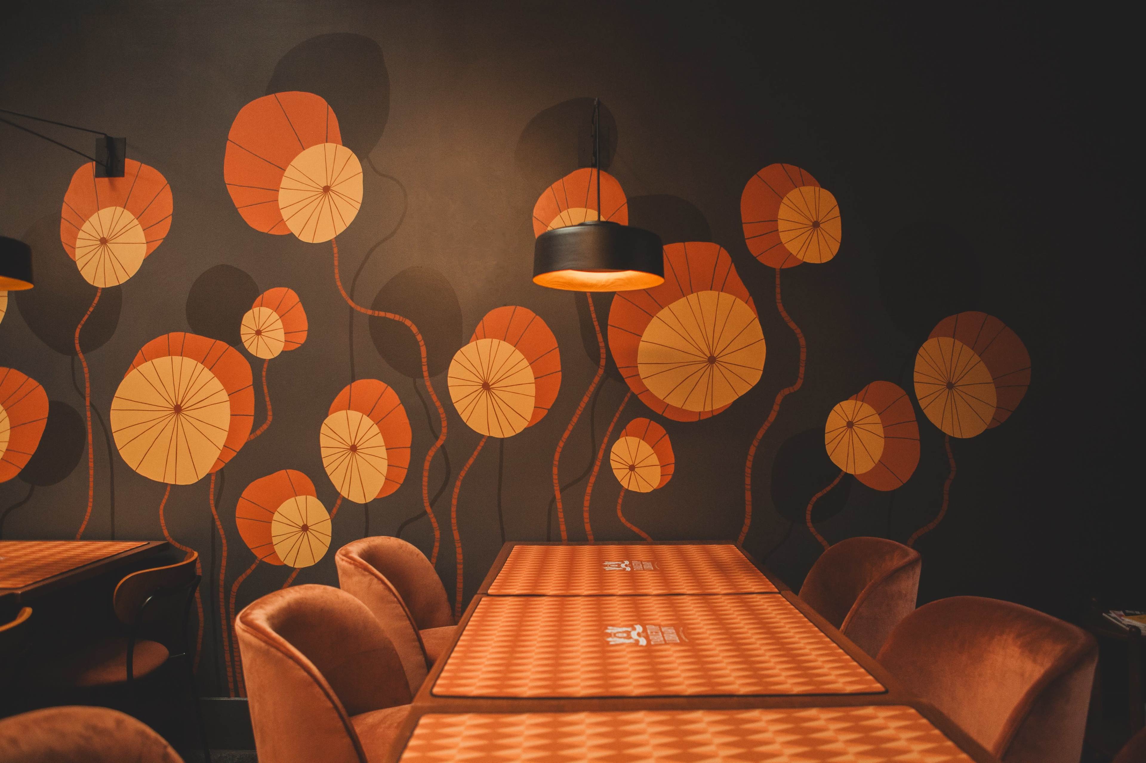 The image shows a dining area with tables covered in patterned tablecloths and a wall adorned with abstract floral designs in warm colors.