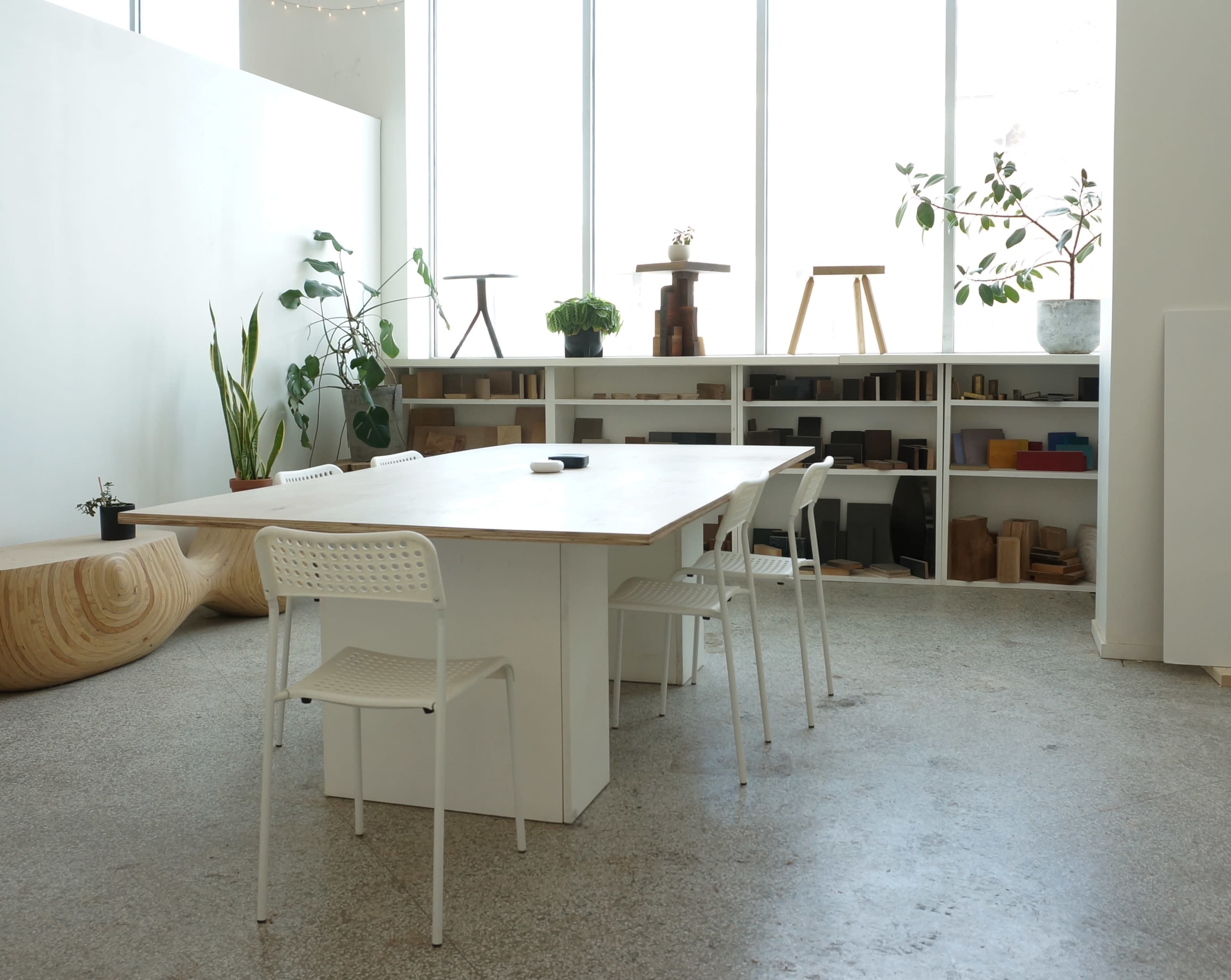 The image shows a minimalist meeting room featuring a large wooden table surrounded by four white chairs, with plants and shelves displaying various wooden objects in the background.