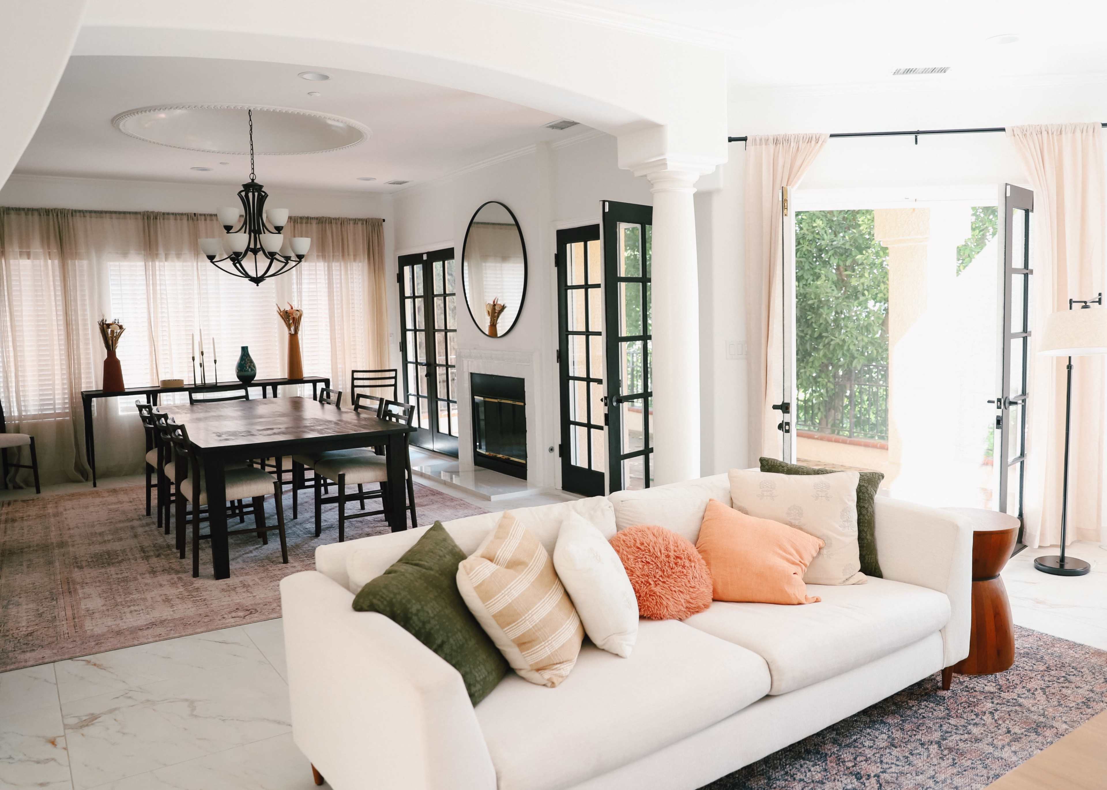 A modern living room features a white sofa with decorative pillows, a dining area with a large table, and large windows with sheer curtains.