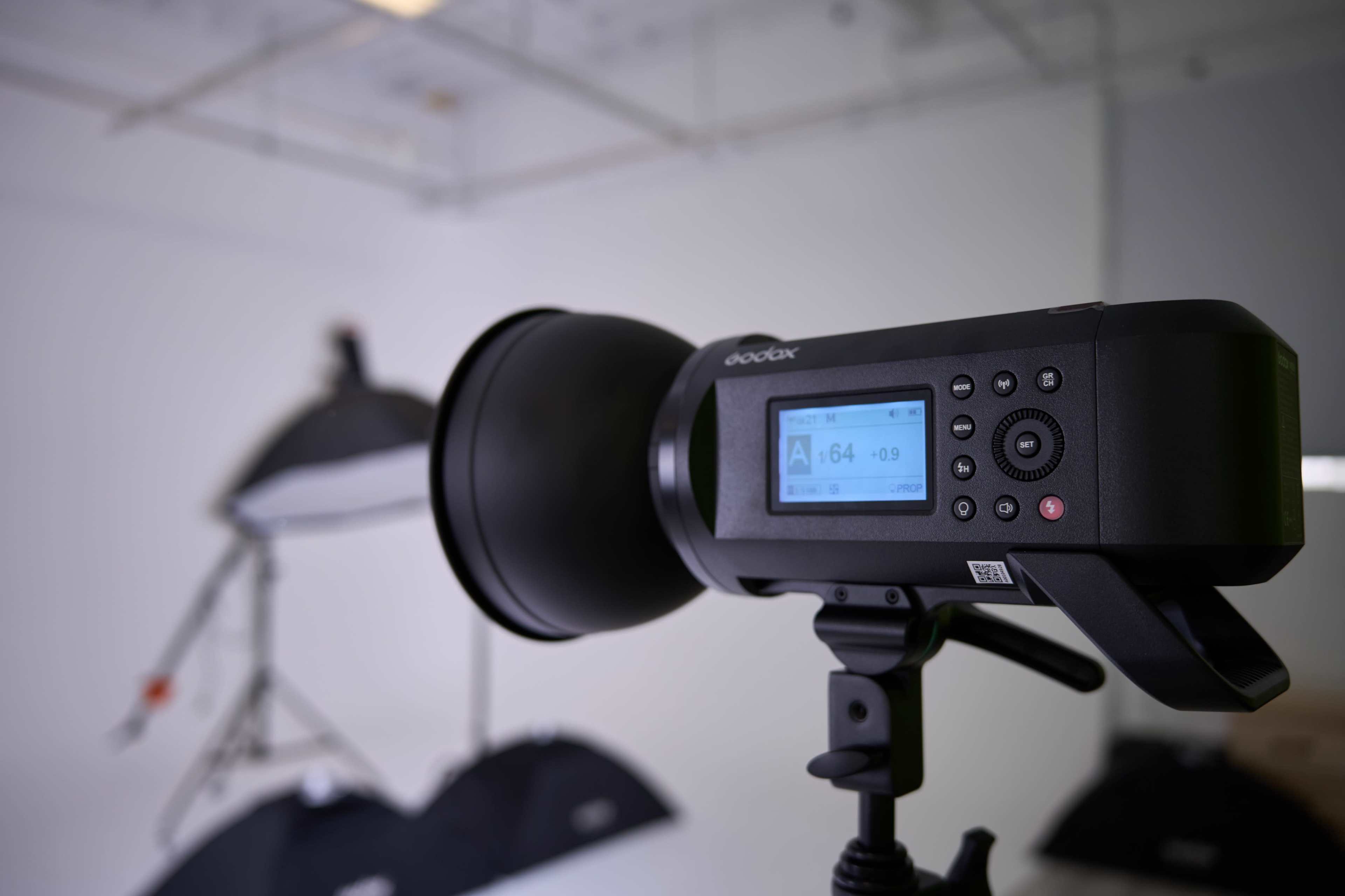 A studio strobe light is mounted on a stand, with its digital display showing settings, and softboxes are visible in the background.