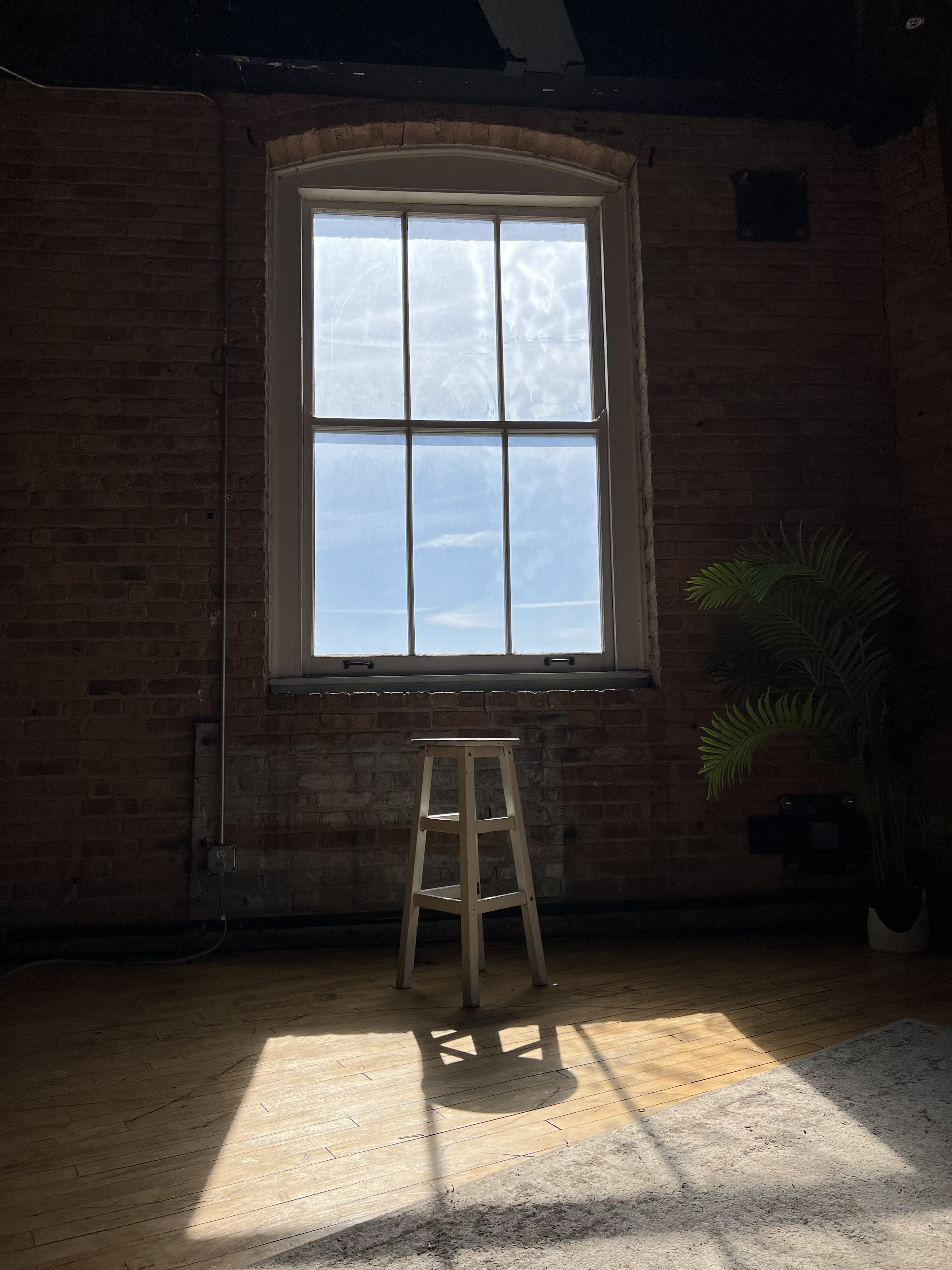 A wooden stool stands in a sunlit room beside a large window, casting shadows on the wooden floor.