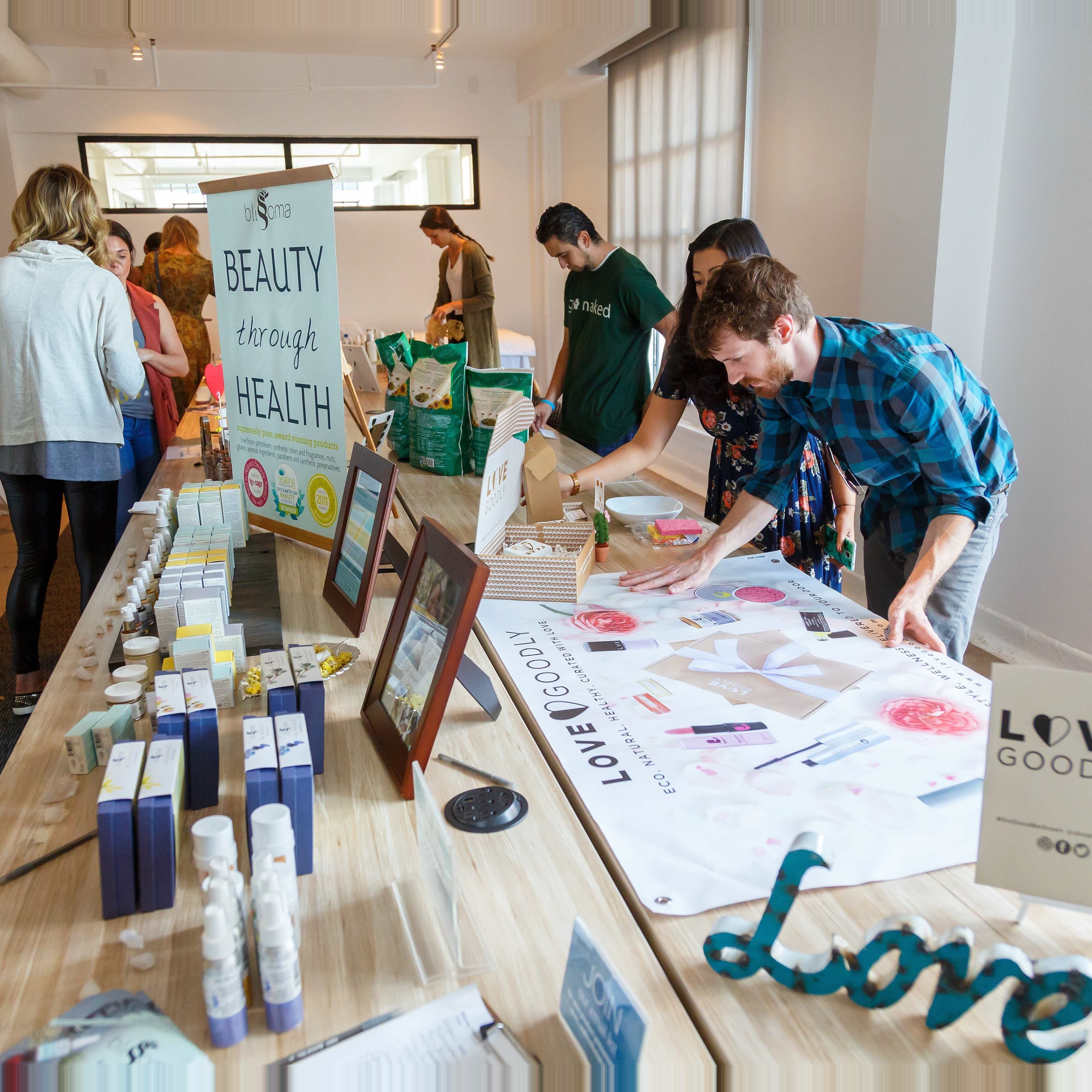 The image shows a group of people interacting at a health and wellness event with a long table featuring various products and informational materials.