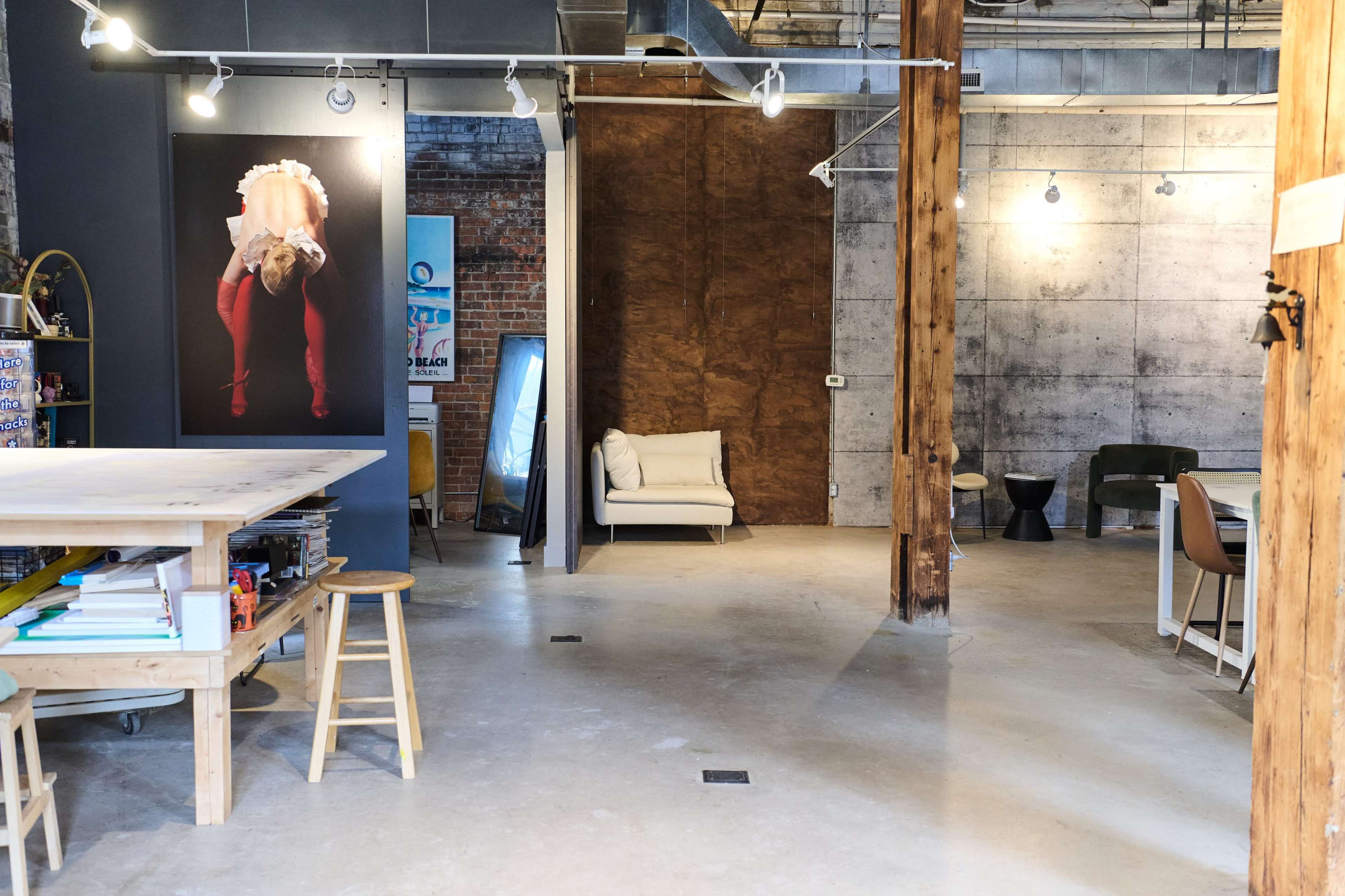The image depicts an industrial-style studio space with wooden beams, a large table, and a minimalist seating area featuring a white couch against a concrete wall.