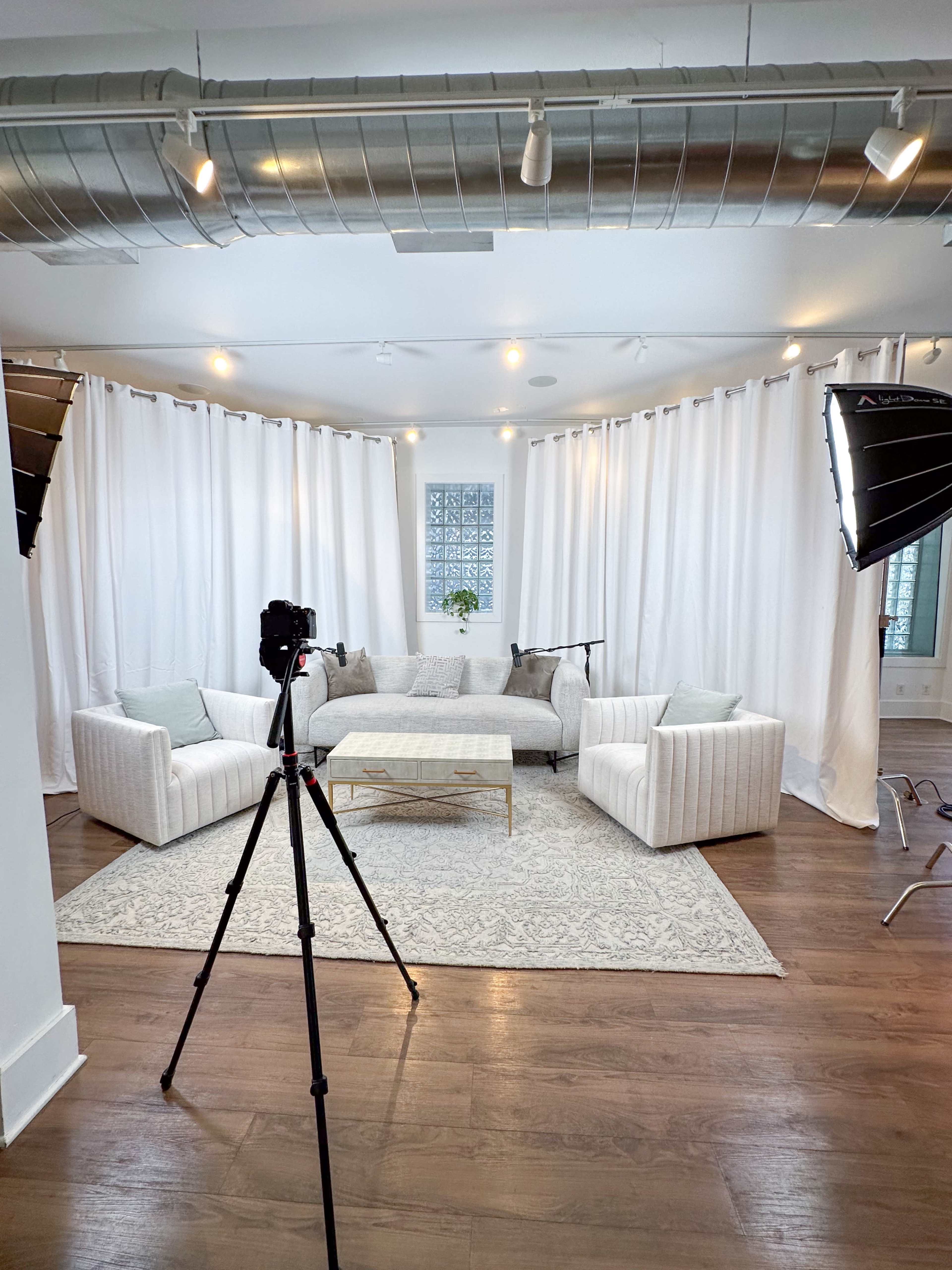 A spacious room features a white sectional sofa, a coffee table on a patterned rug, and a camera set up on a tripod, surrounded by sheer curtains and studio lights.