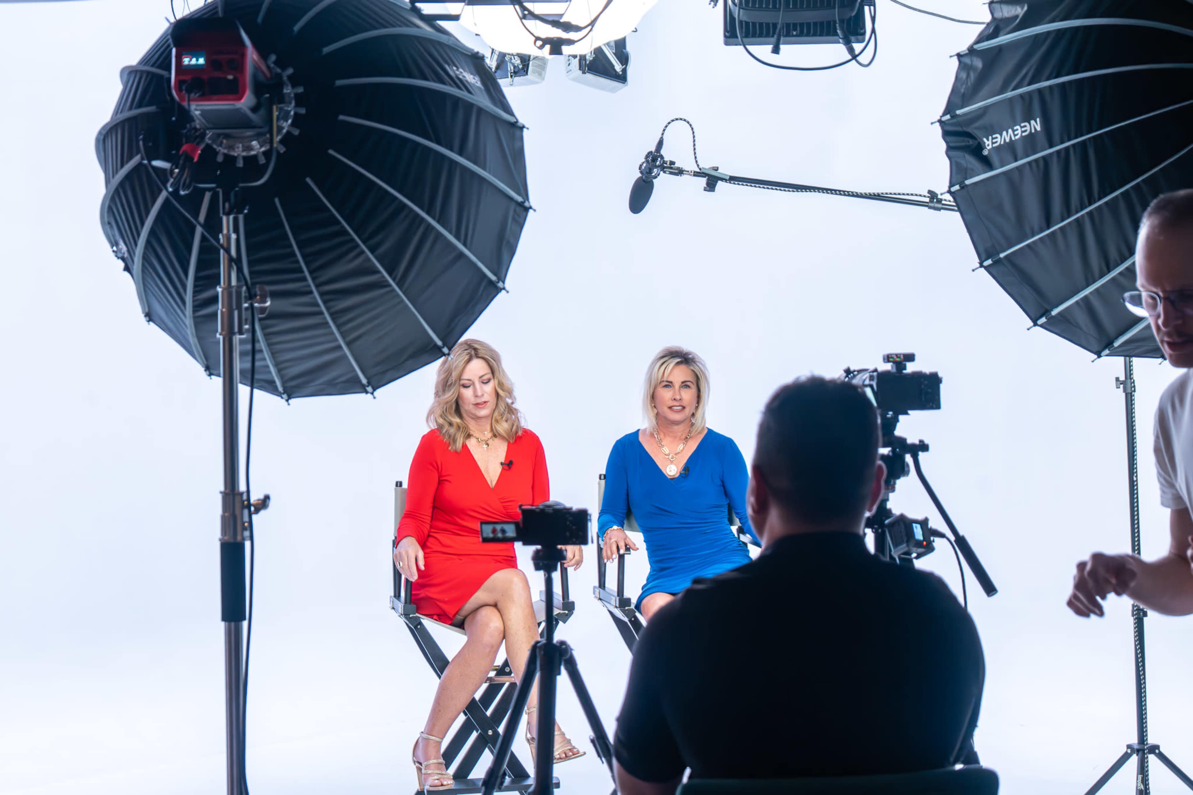Two women sit in director's chairs against a white backdrop, flanked by large softbox lights, while a person operates a camera in front of them.
