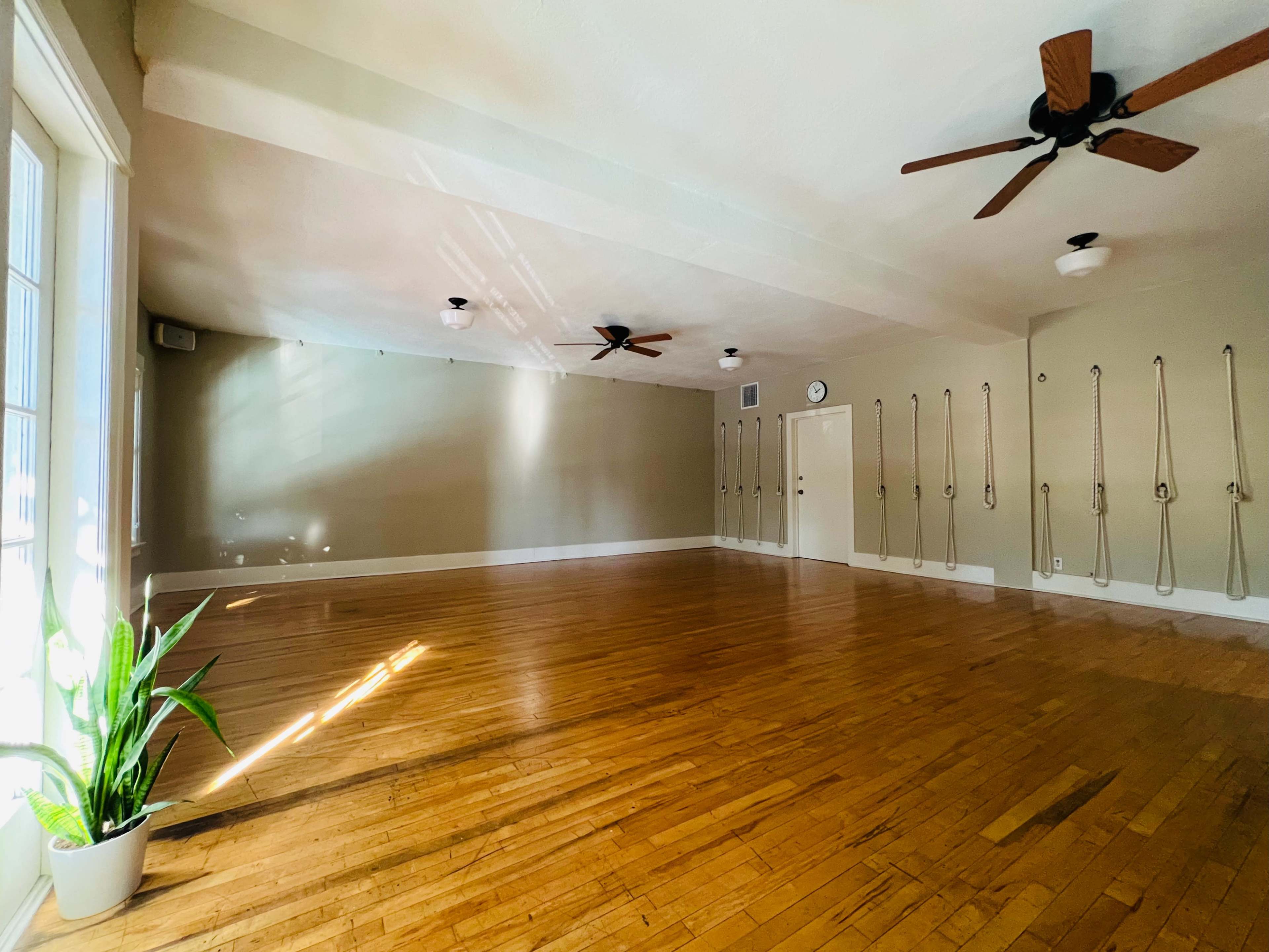 A spacious yoga studio with wooden floors, wall ropes, and ceiling fans, illuminated by natural light from large windows.