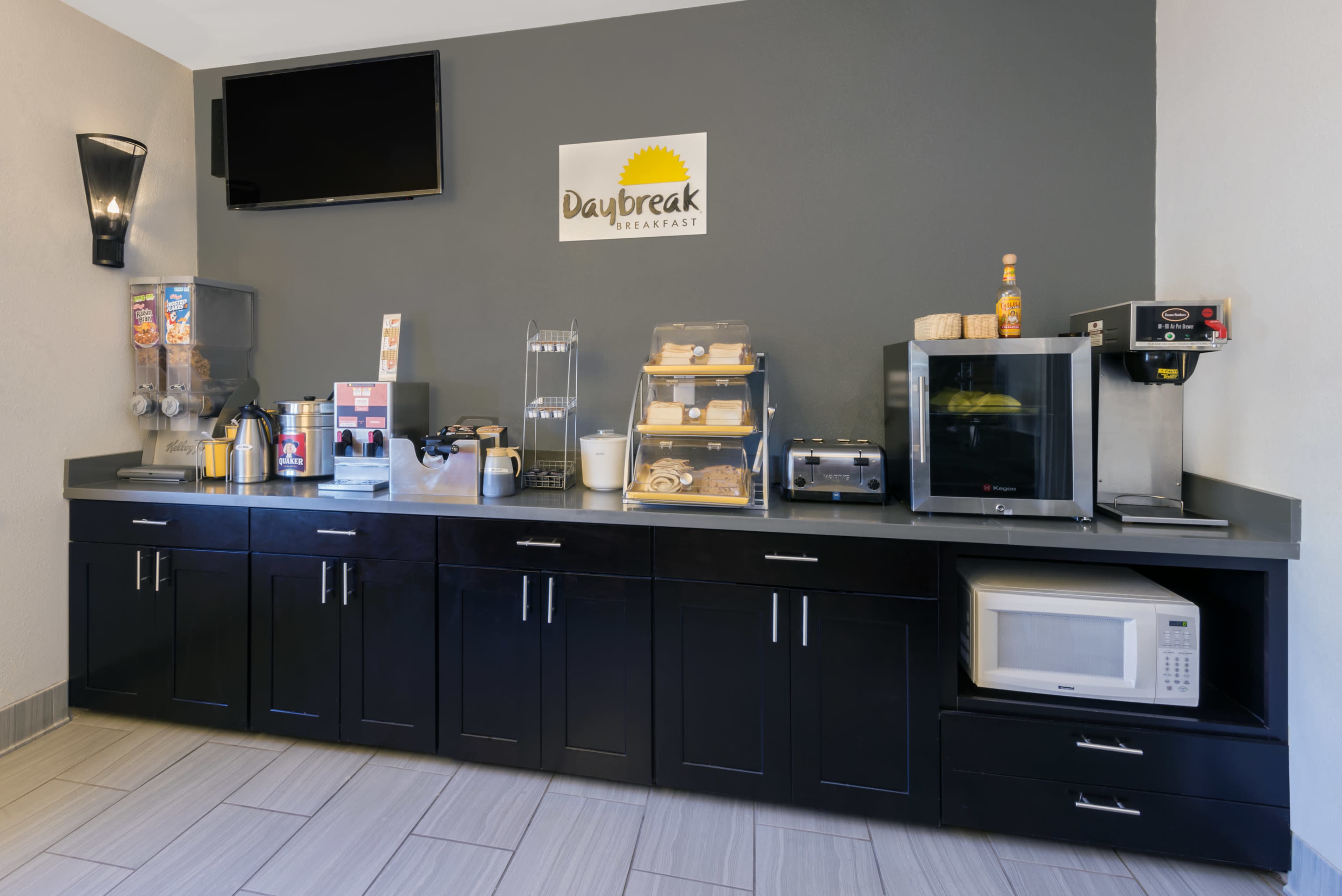 A breakfast area features a countertop with various appliances, including a coffee machine, toaster, microwave, and food display cases, against a gray wall.