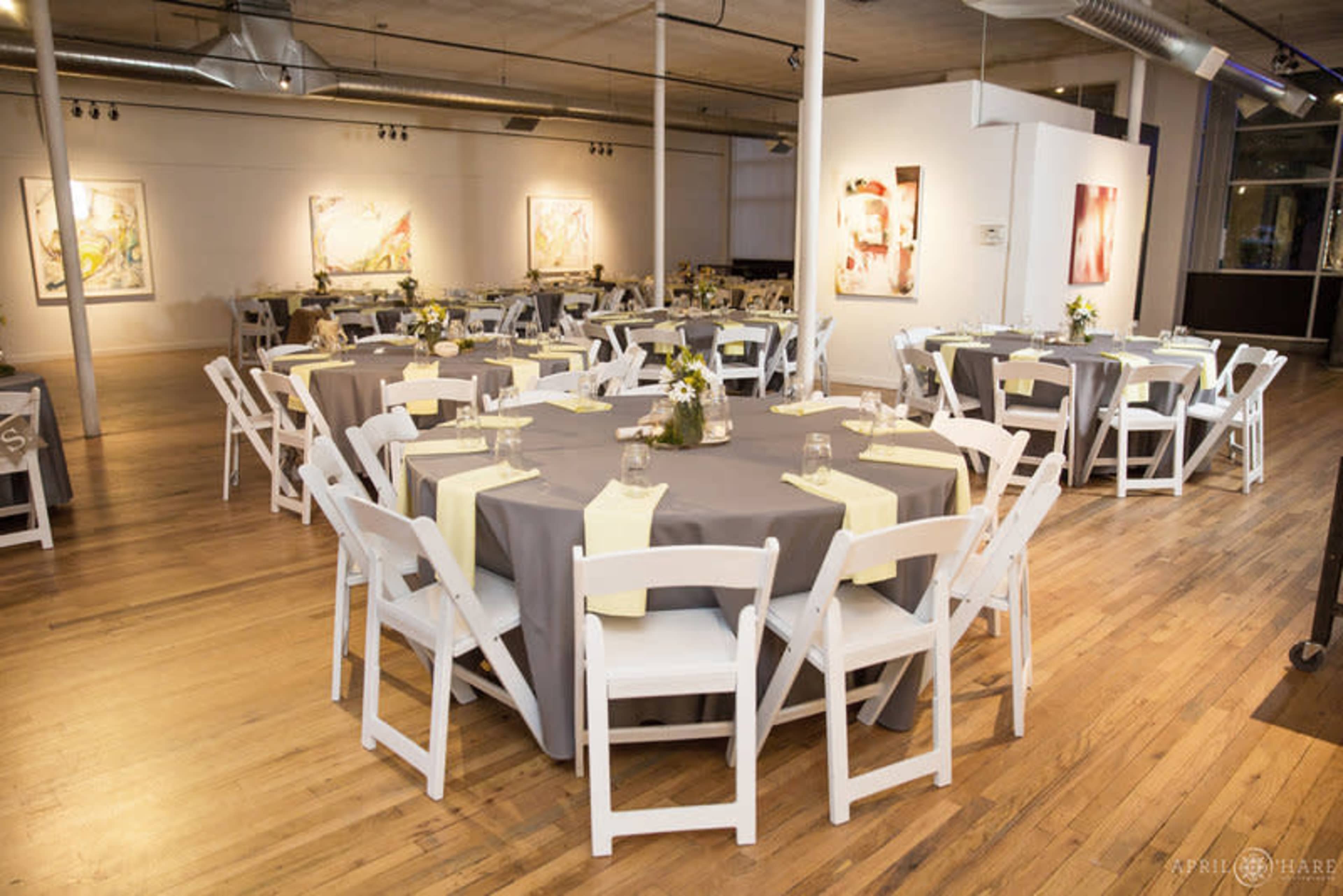 The image shows a banquet hall set up for an event, featuring round tables covered with gray tablecloths and adorned with yellow napkins, surrounded by white chairs.