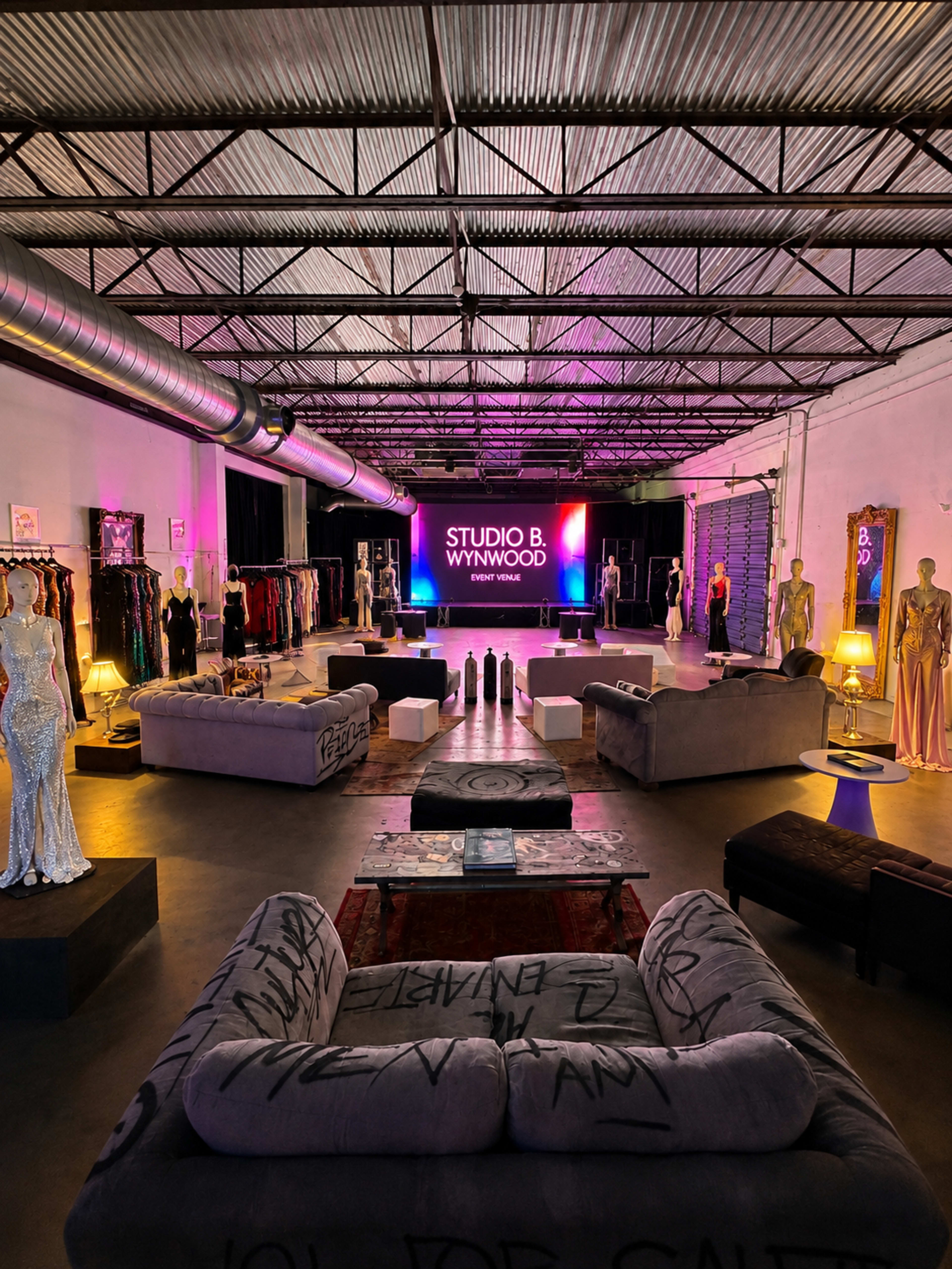 The image shows a spacious studio with a high ceiling, featuring stylish furniture, mannequins displaying clothing, and a large screen that reads "STUDIO B WYNWOOD."