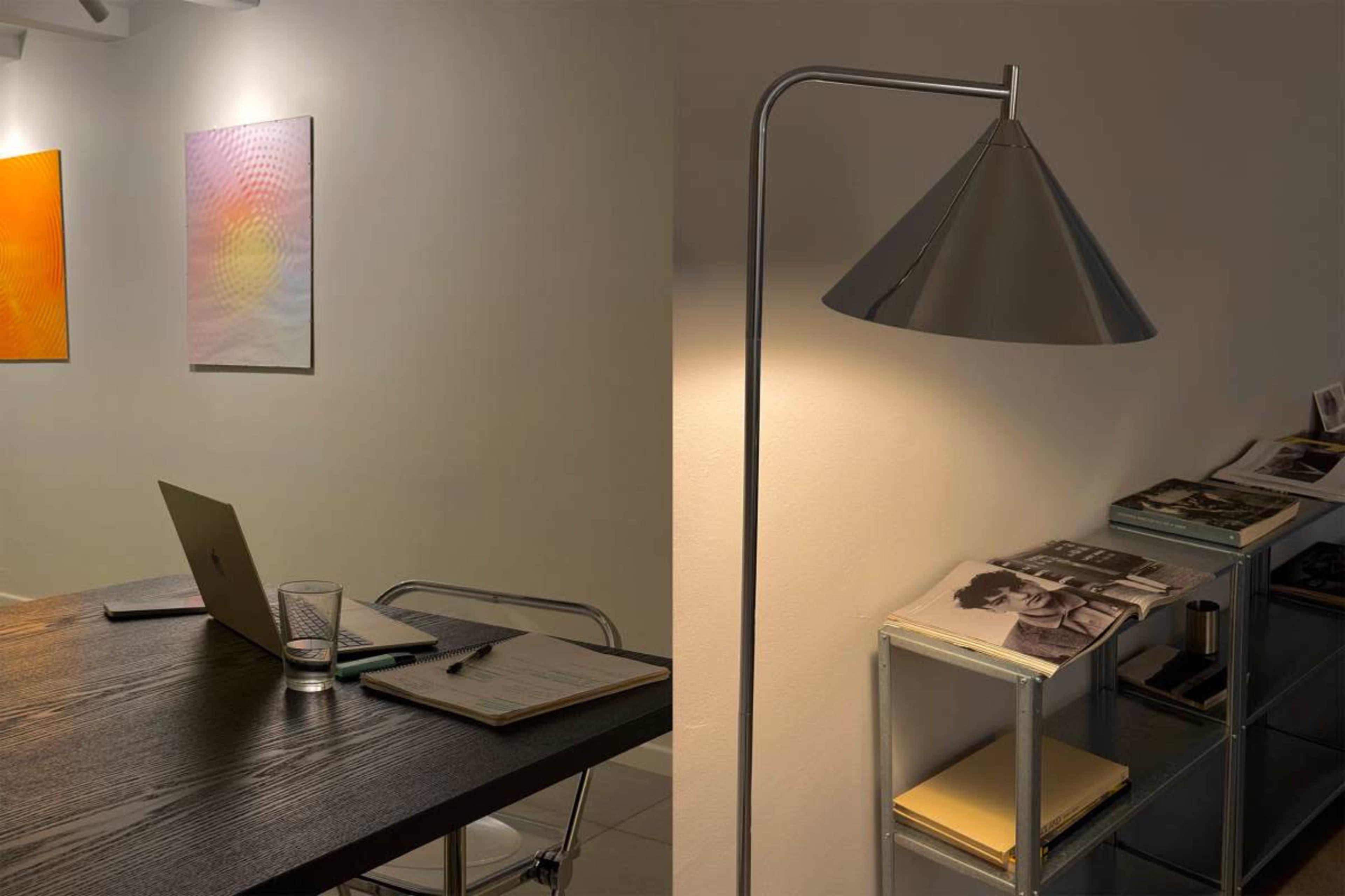 The image shows a minimalist workspace featuring a wooden table with a laptop, a glass of water, a notepad, and a contemporary lamp beside a shelf with stacked magazines and books.