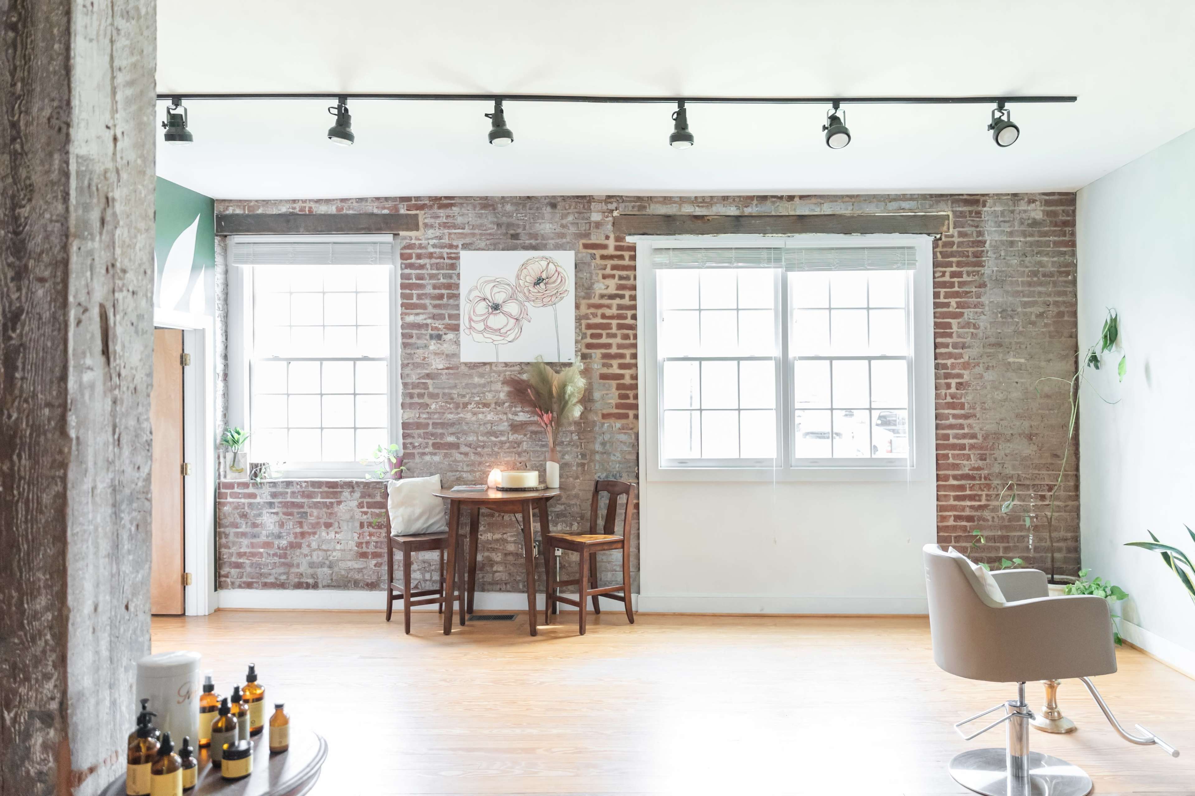 A cozy room features exposed brick walls, a small table with two chairs, and large windows allowing natural light.