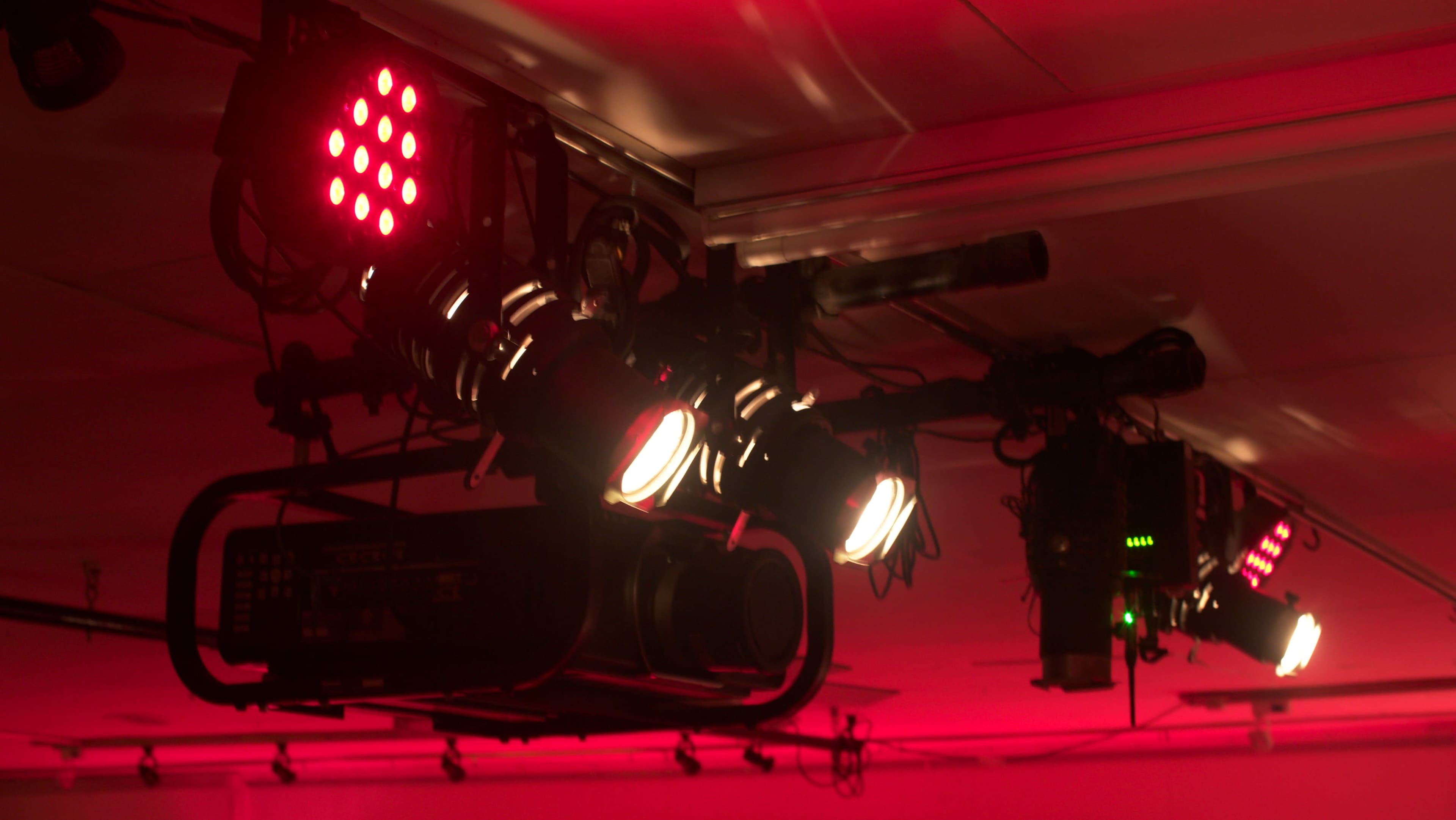 A series of studio lights and a projector hang from a ceiling, illuminated by red lighting.