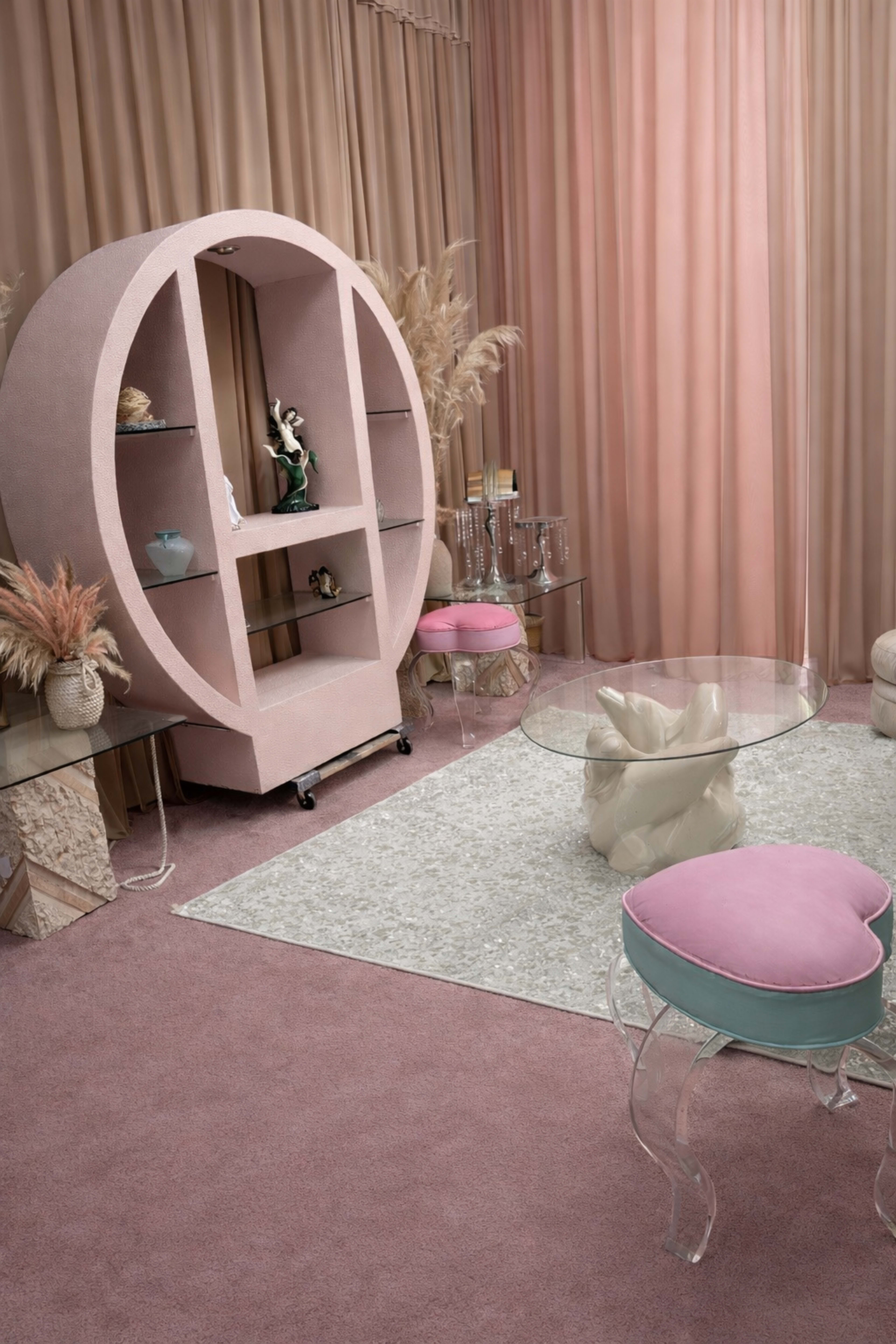The scene features a designed interior with a circular, pink bookshelf, a glass table, heart-shaped transparent stools, and soft, neutral-toned curtains.