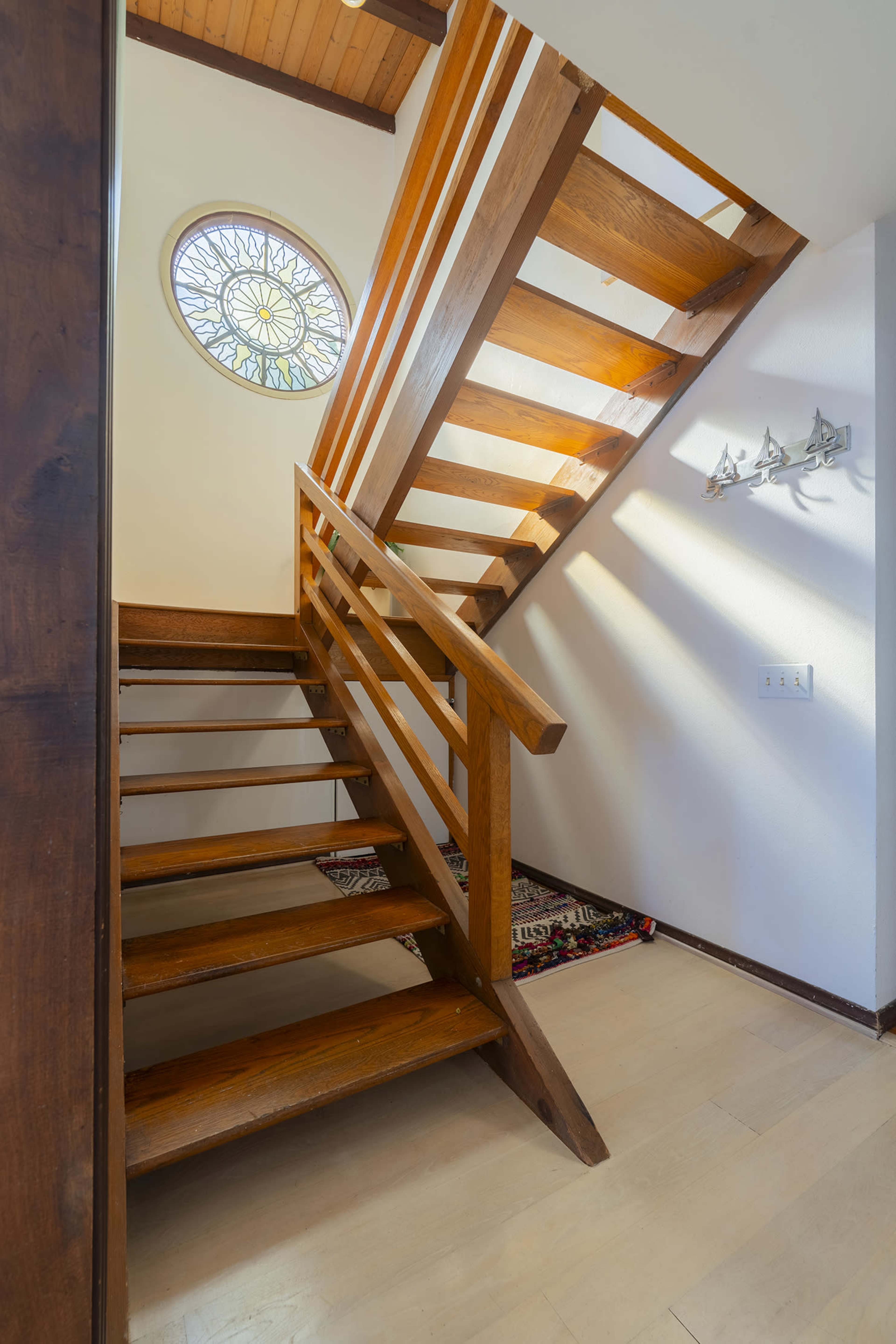 The image shows a wooden staircase with a circular stained glass window above, leading to an upper level in a well-lit interior space.