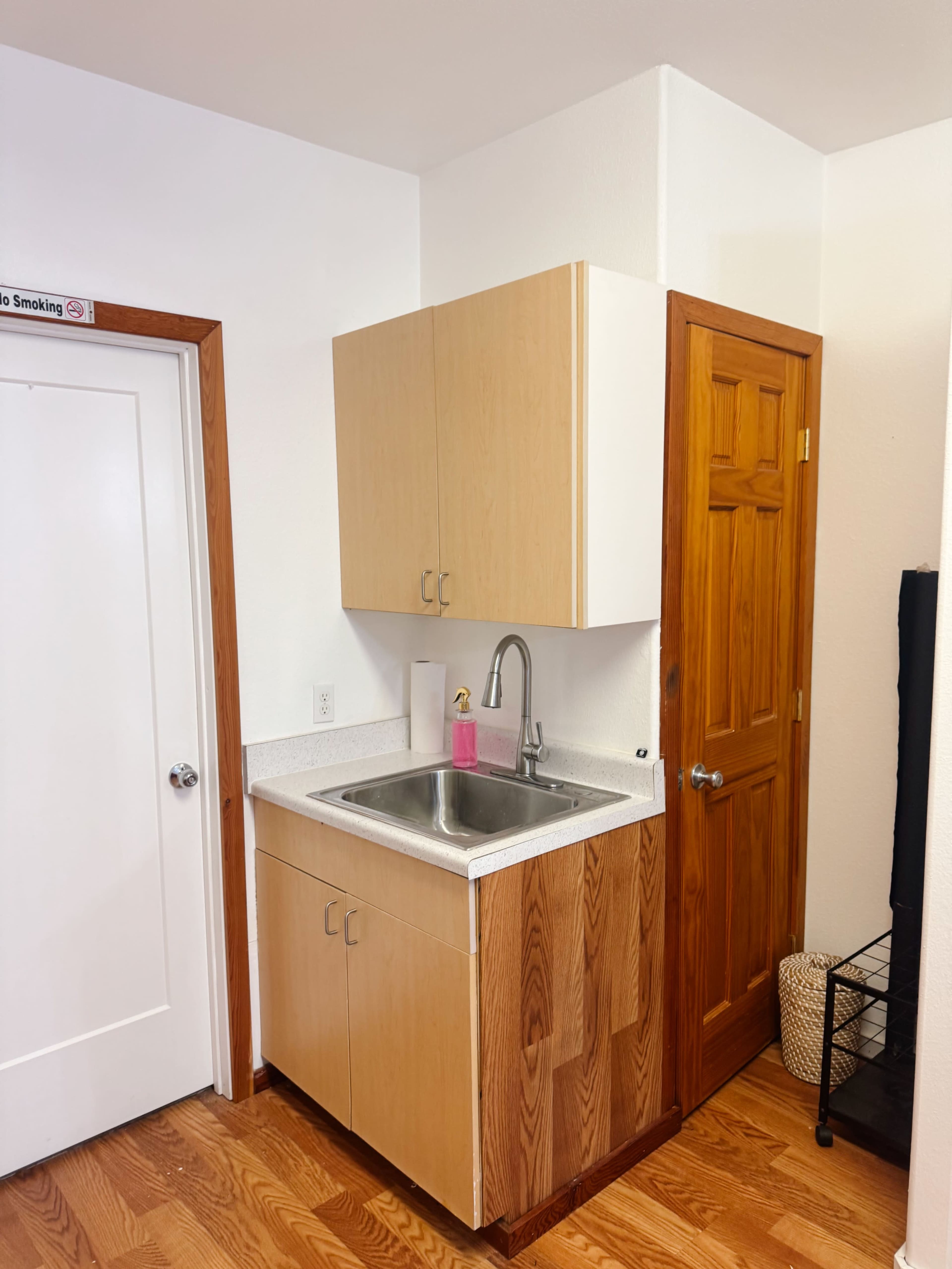 The image shows a small kitchenette area with a sink and wooden cabinets, located next to a white door.
