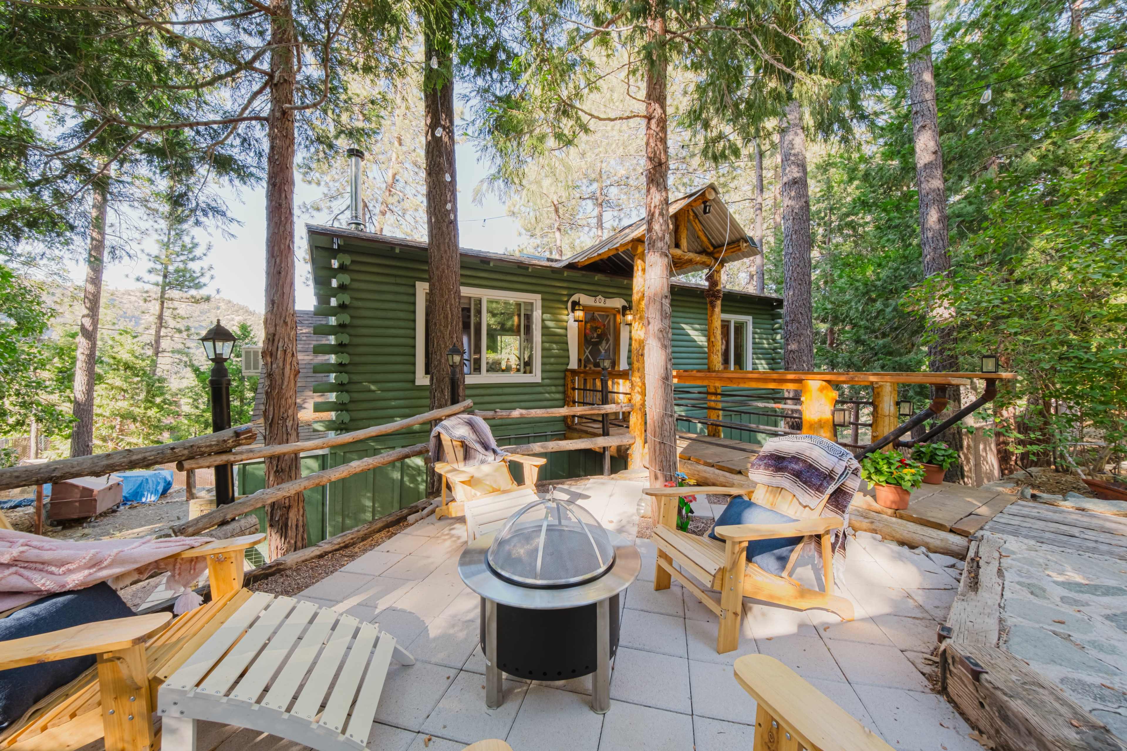 A wooden cabin nestled among tall trees features a patio area with chairs and a fire pit.