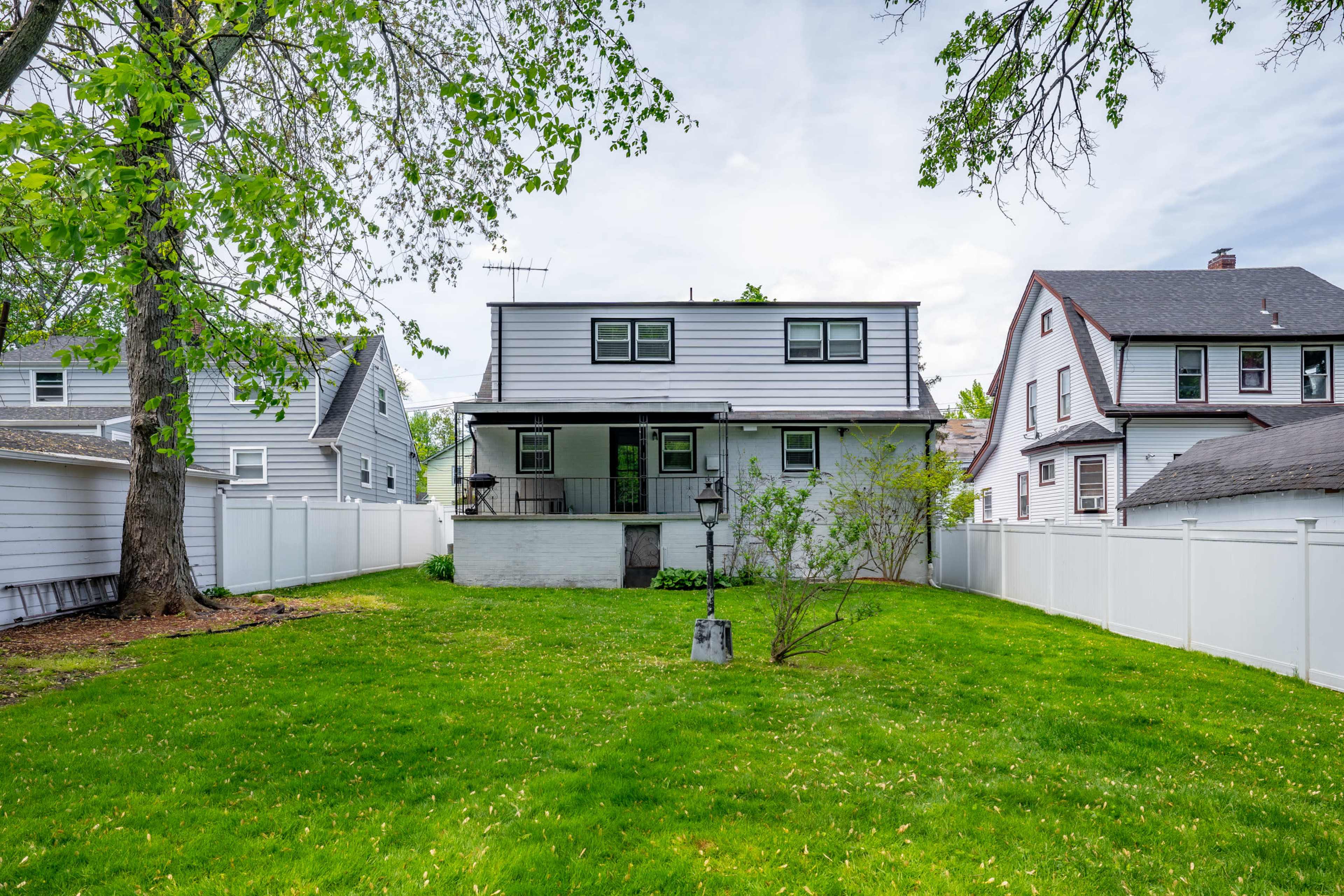 A two-story house with gray siding is situated in a backyard surrounded by a white privacy fence and grass.
