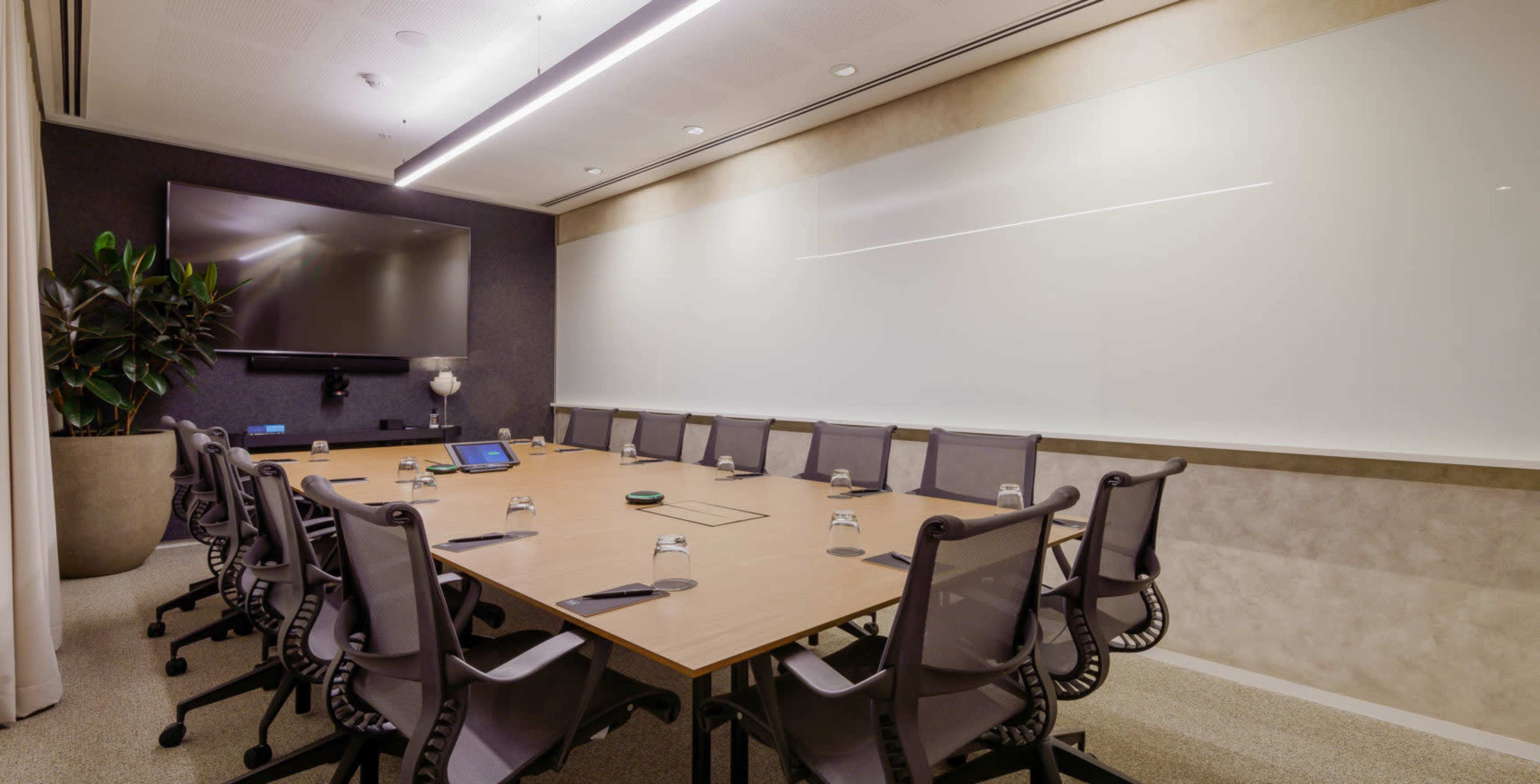 A modern conference room features a large table surrounded by ergonomic chairs, a wall-mounted television, and a whiteboard.