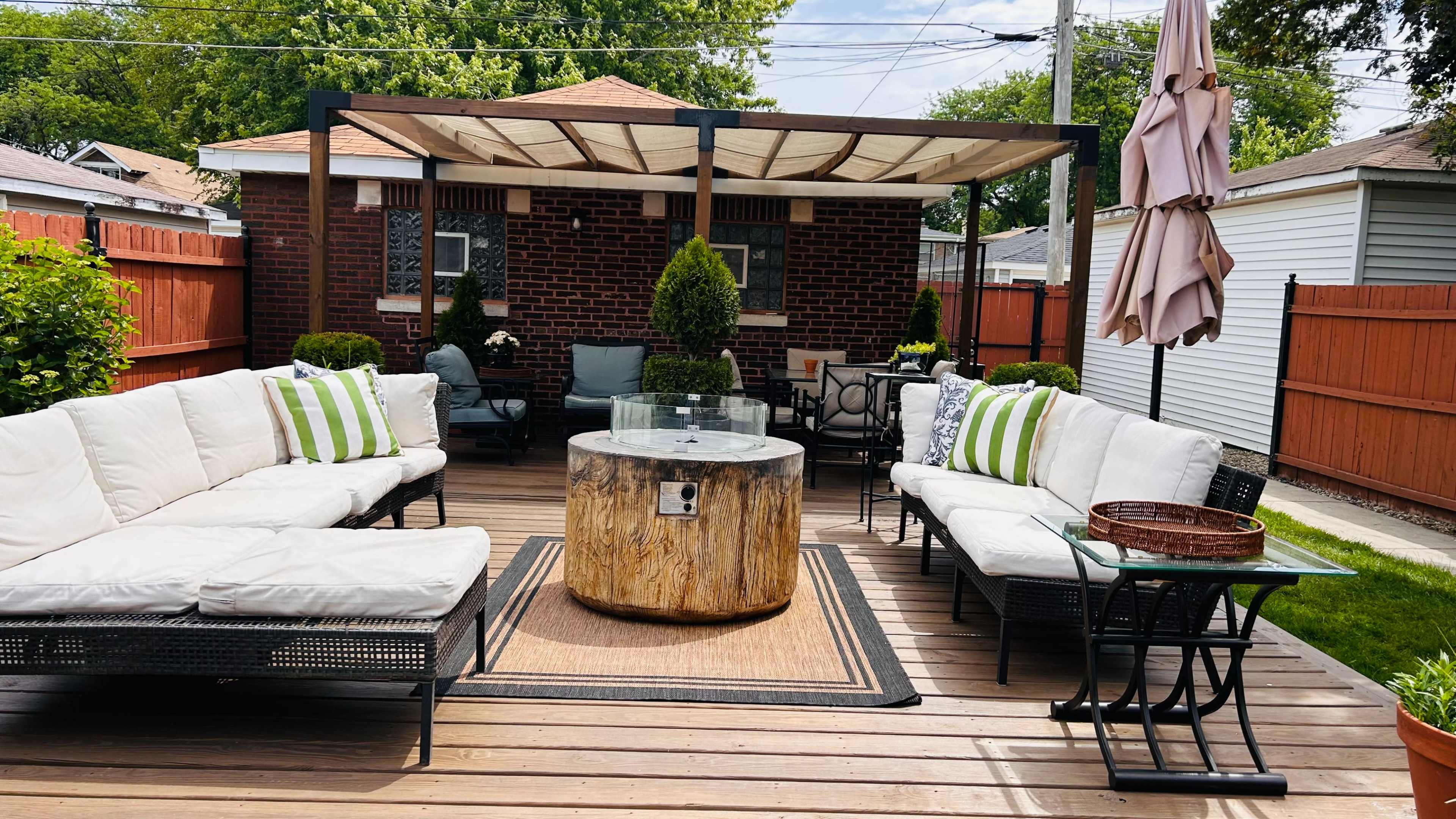The image shows a backyard patio area with a wooden deck, multiple seating arrangements, a round table made from a tree stump, and a pergola overhead.
