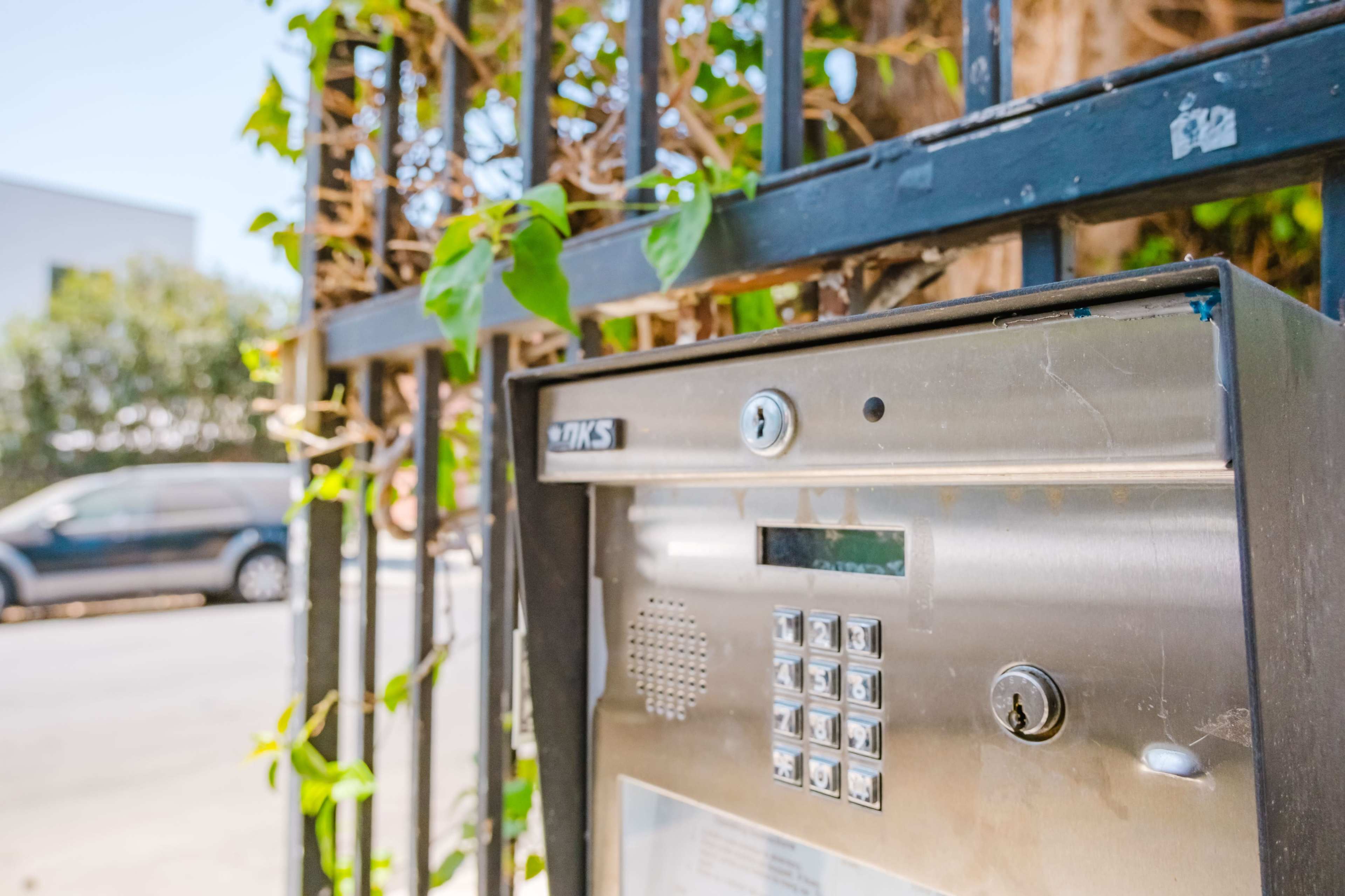 The image shows a metal intercom system mounted on a black wrought iron gate surrounded by leafy green vines.