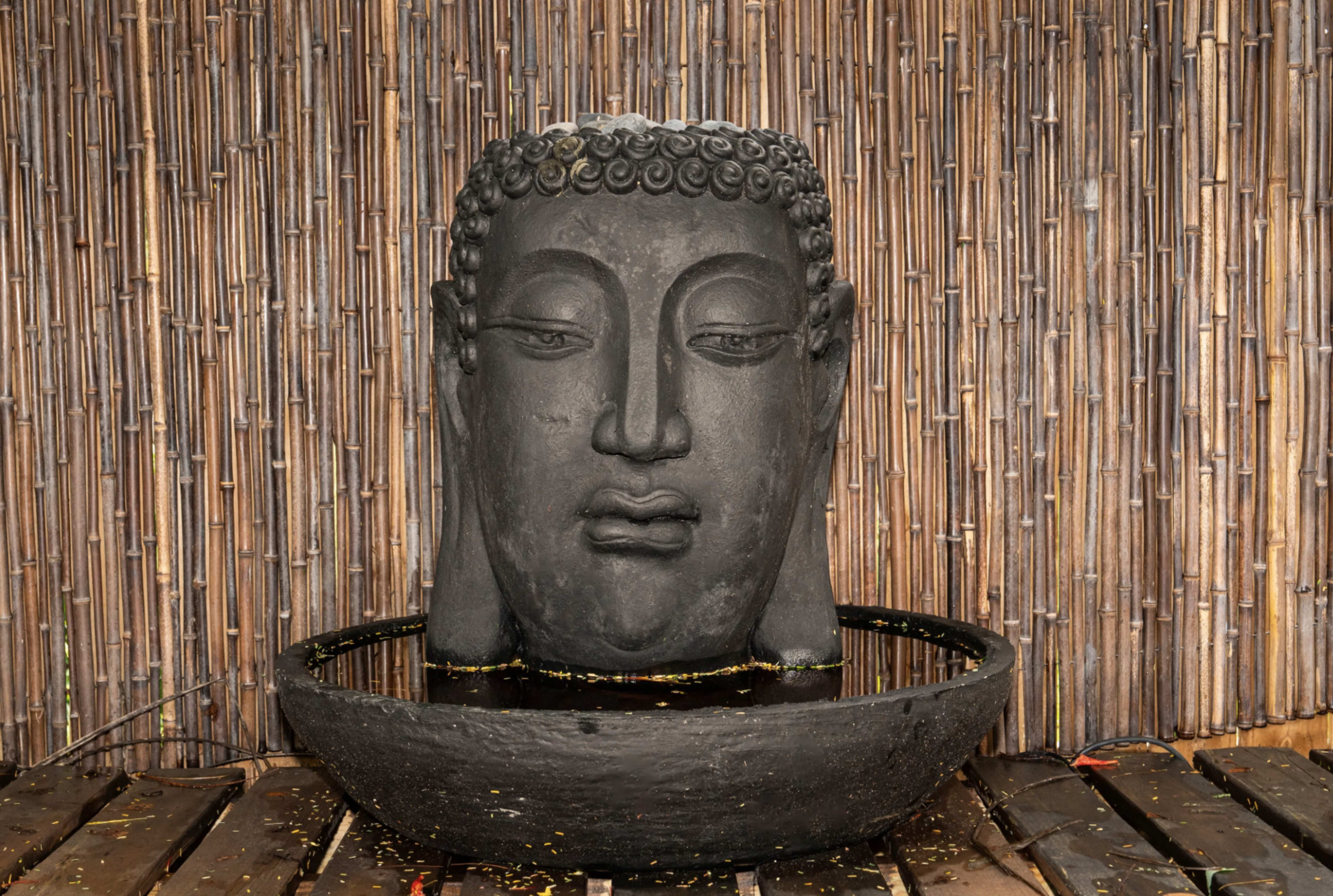 A large black stone Buddha head is mounted in a circular basin, surrounded by a bamboo backdrop.