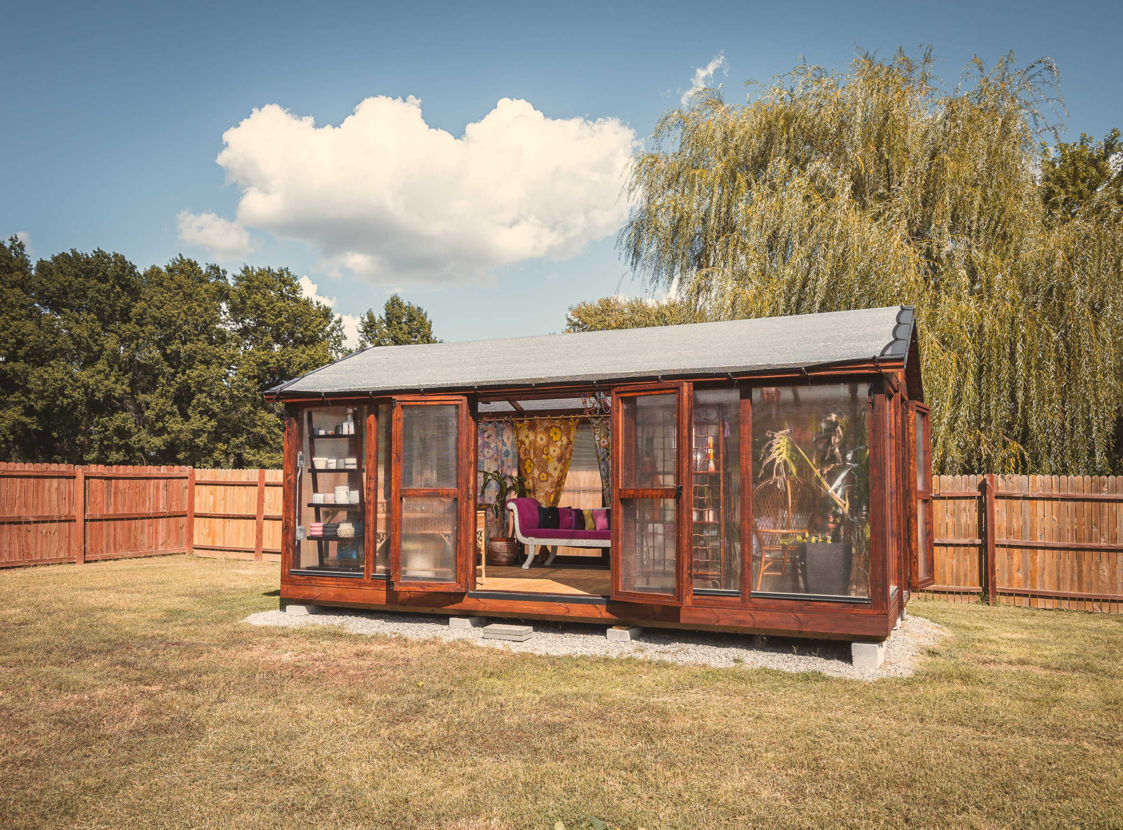 A wooden garden shed with glass walls and a metal roof sits in a grassy yard surrounded by a wooden fence and trees.