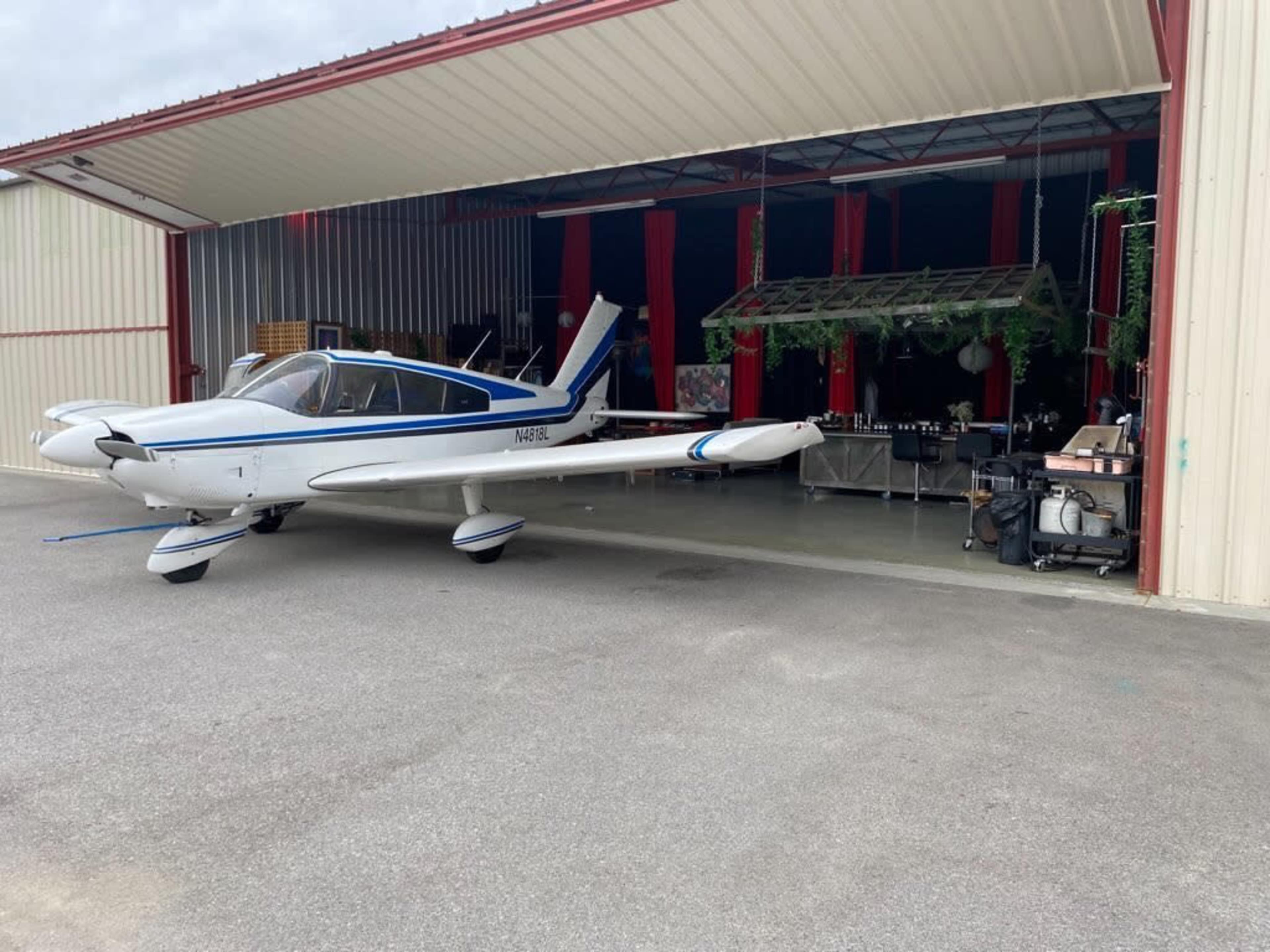A small white and blue aircraft is parked in front of a hangar with its doors open, revealing various equipment and furniture inside.