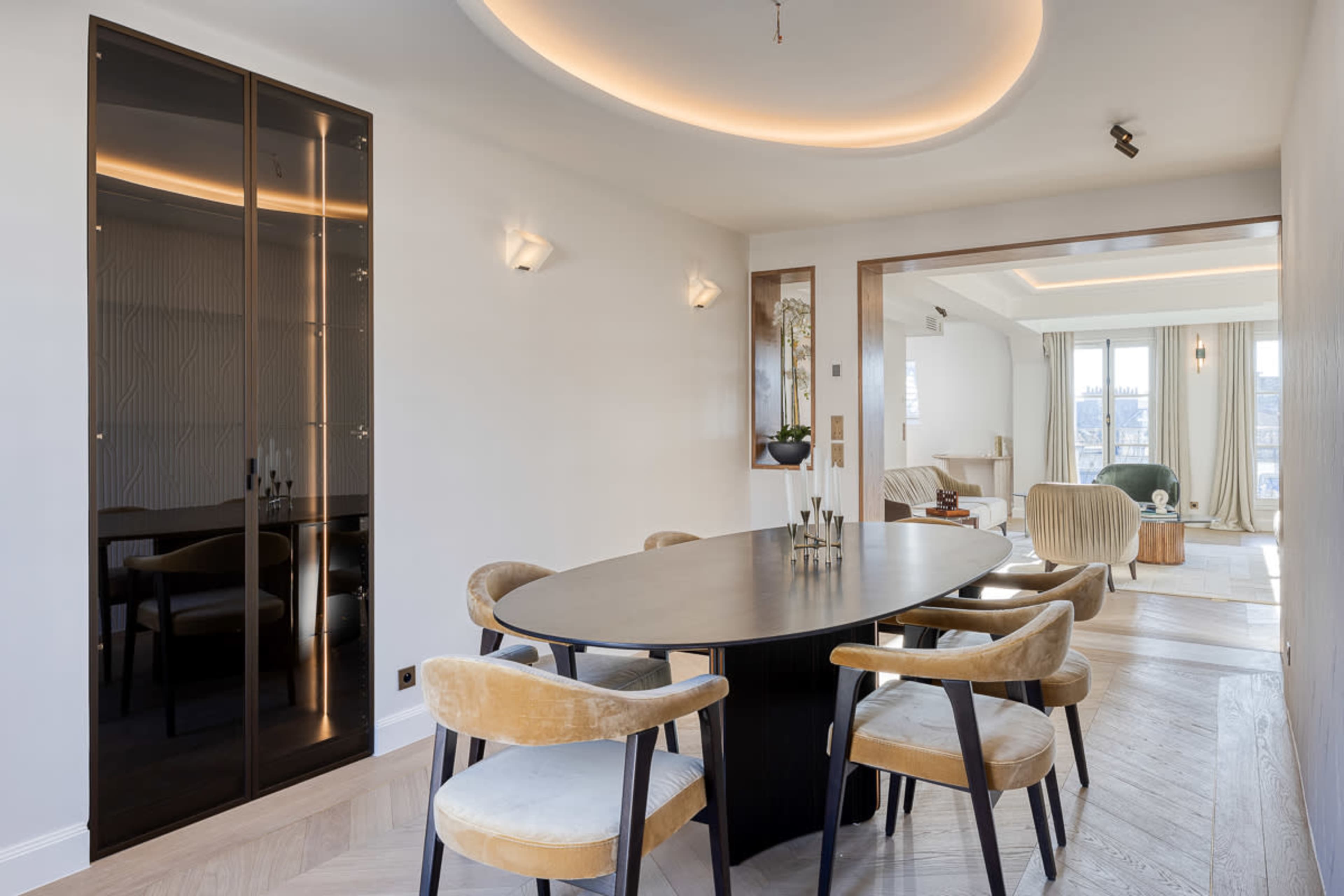 The image shows a modern dining area featuring a circular black table surrounded by upholstered chairs, with a stylish wall-mounted cabinet and an open living space in the background.