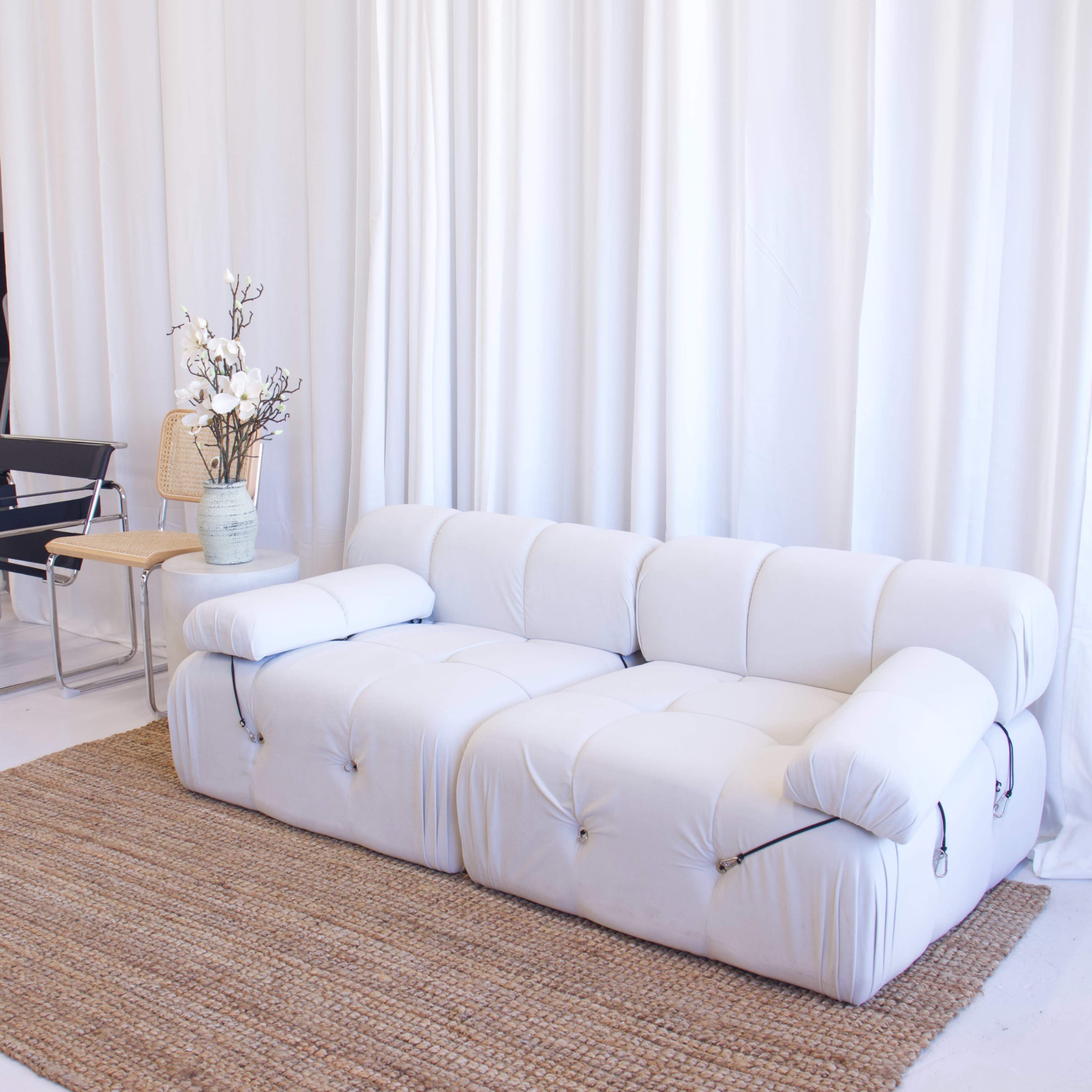 A white, modern sofa with a tufted design is positioned next to a small wooden side table and a decorative vase with flowers, set against a white curtain backdrop.