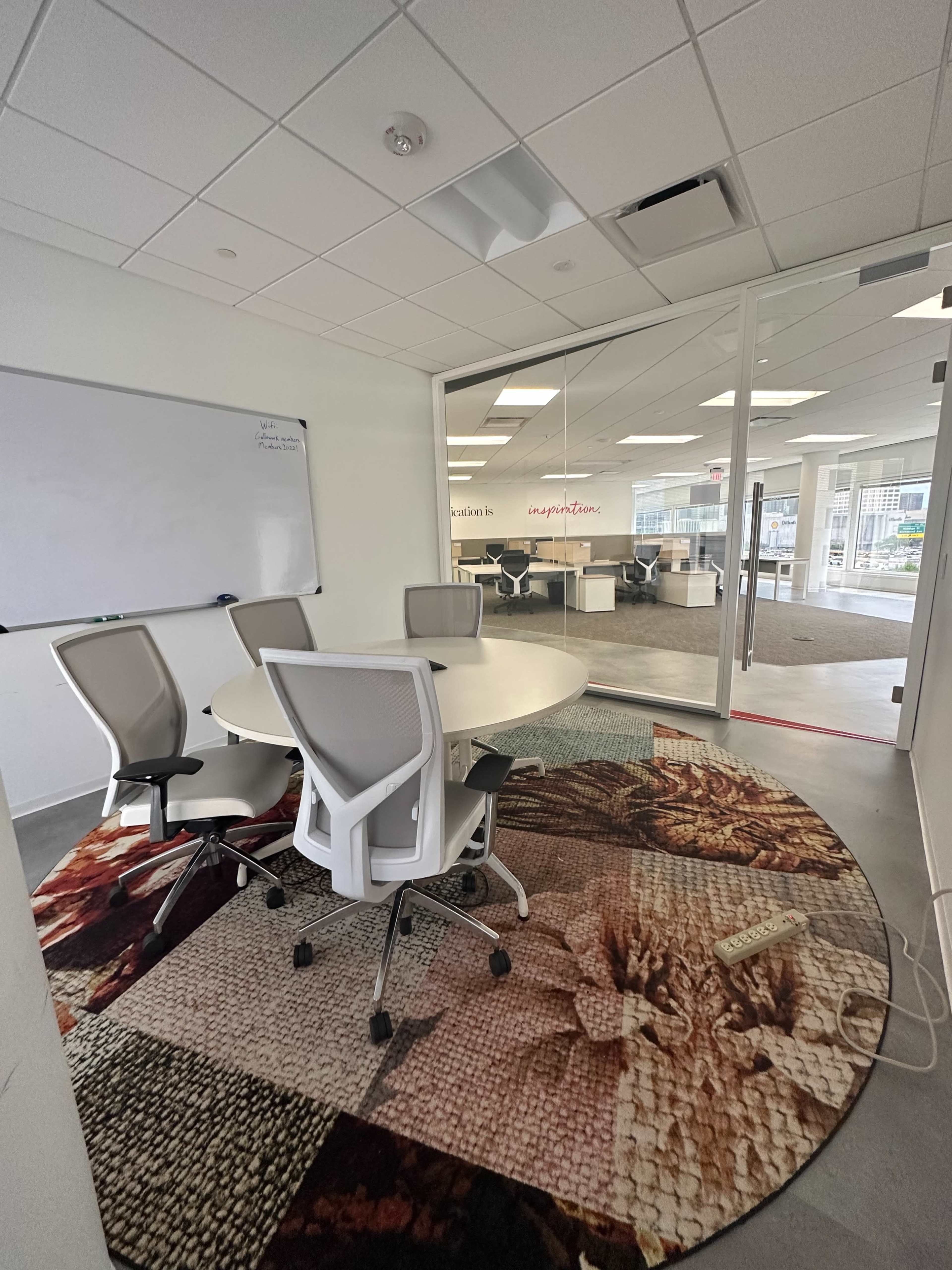 The image shows a modern conference room with a round table, four chairs, a whiteboard, and a decorative rug, enclosed by glass walls.