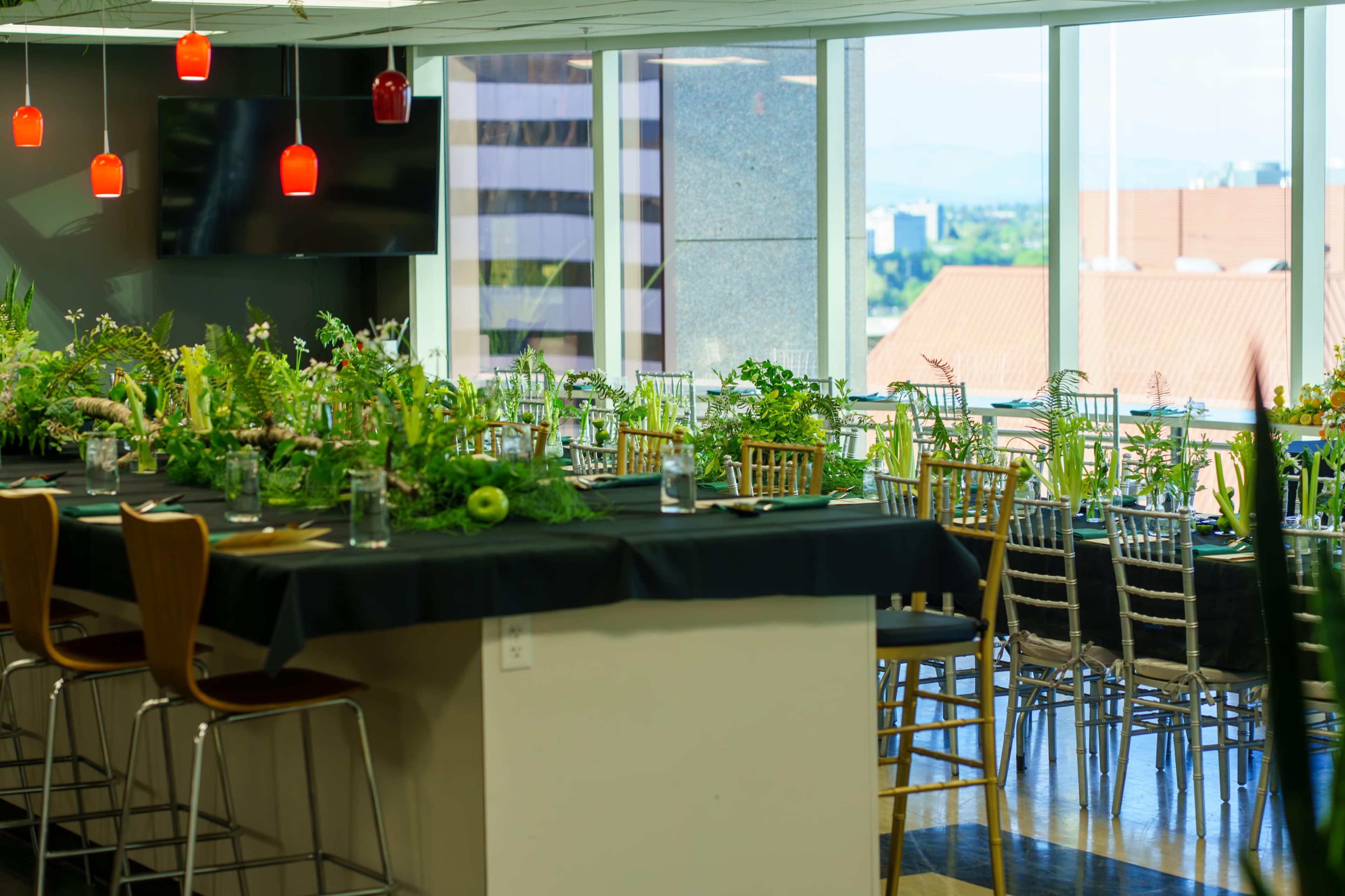 The image shows a modern dining area with large windows, featuring a long table adorned with greenery and floral arrangements, along with bar stools and pendant lighting.