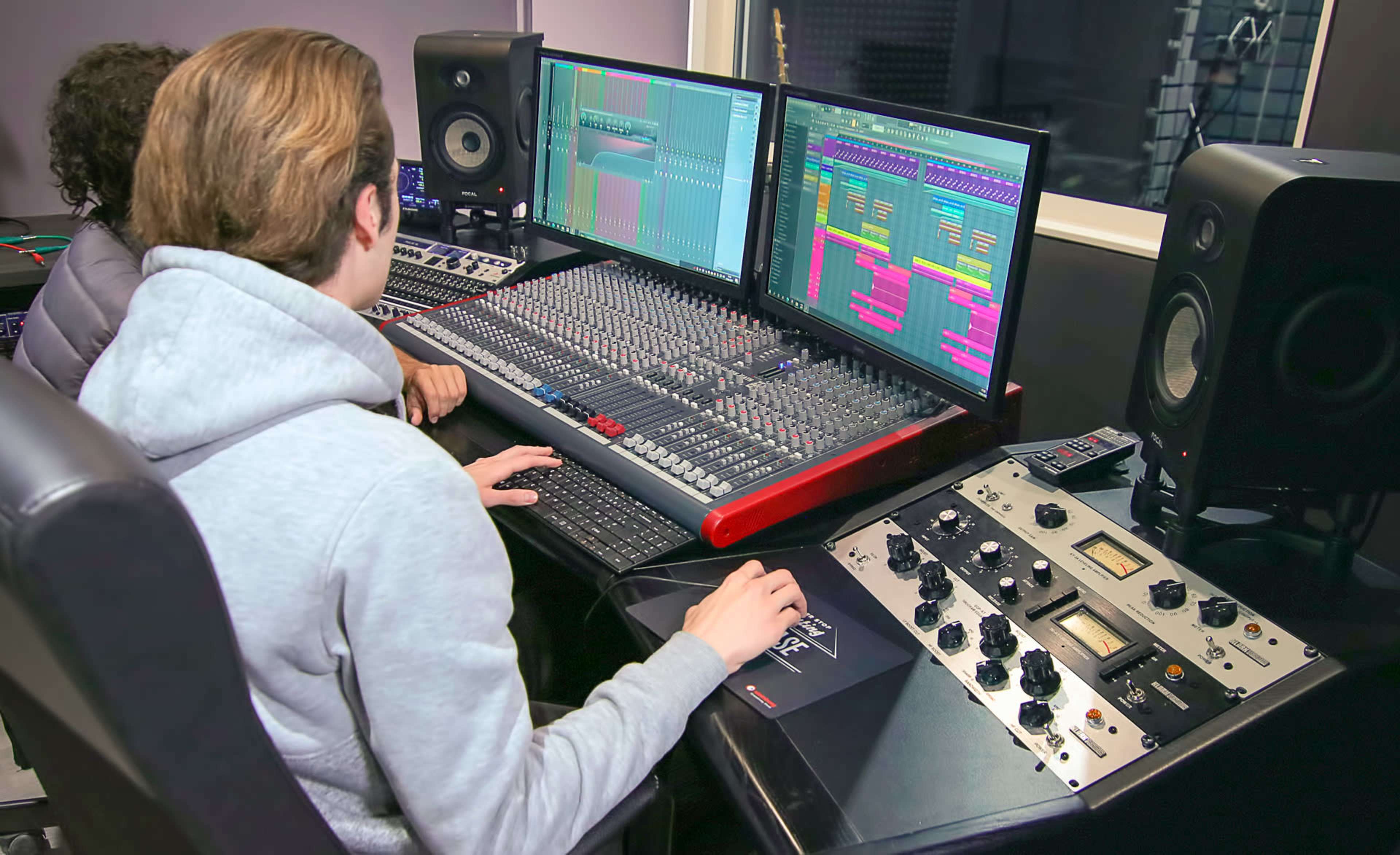 Two individuals are working at a professional audio mixing console illuminated by dual computer monitors displaying music software.