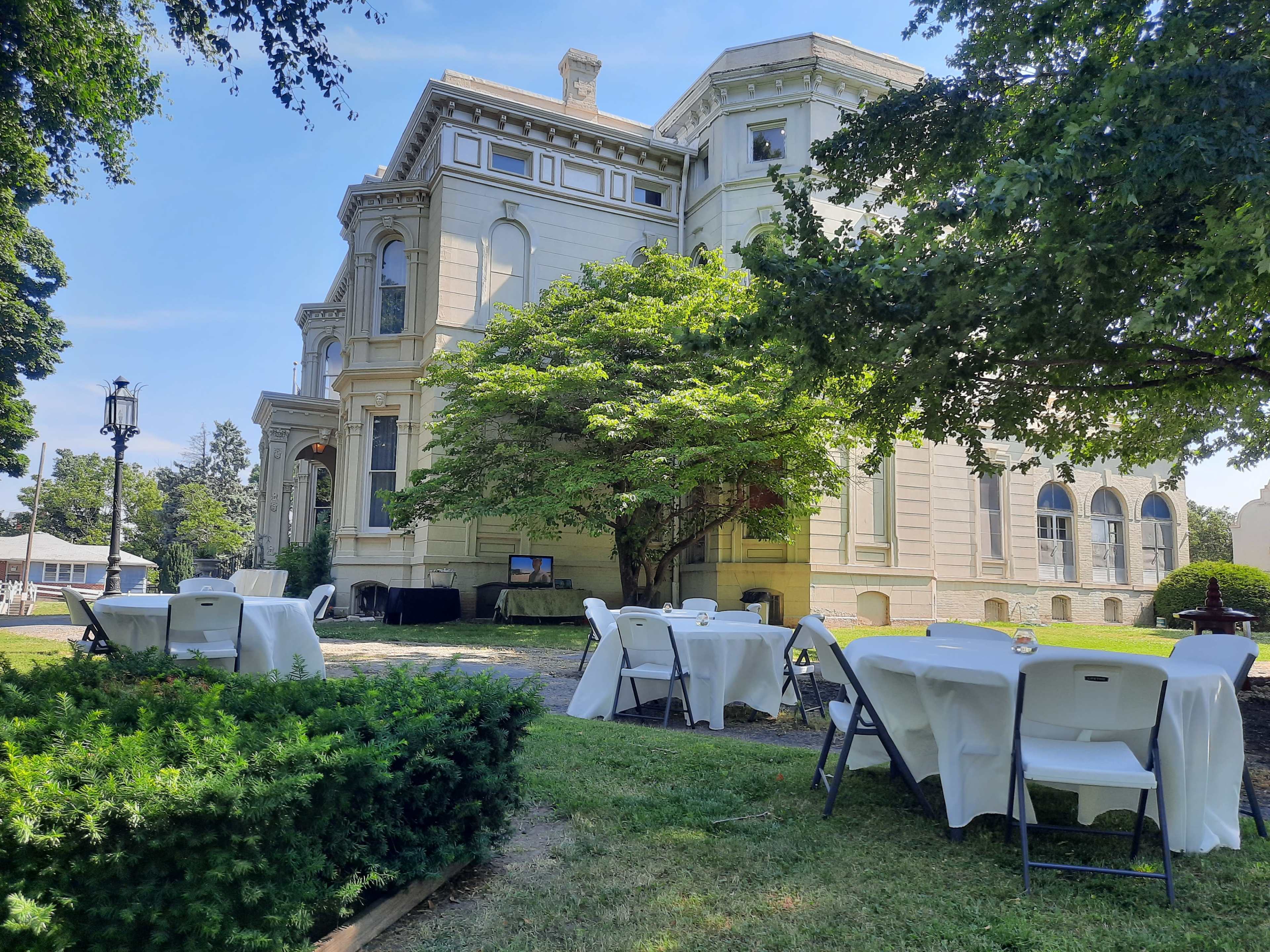 A historic mansion with white walls is surrounded by tables covered in white cloths and a large tree in a grassy area.