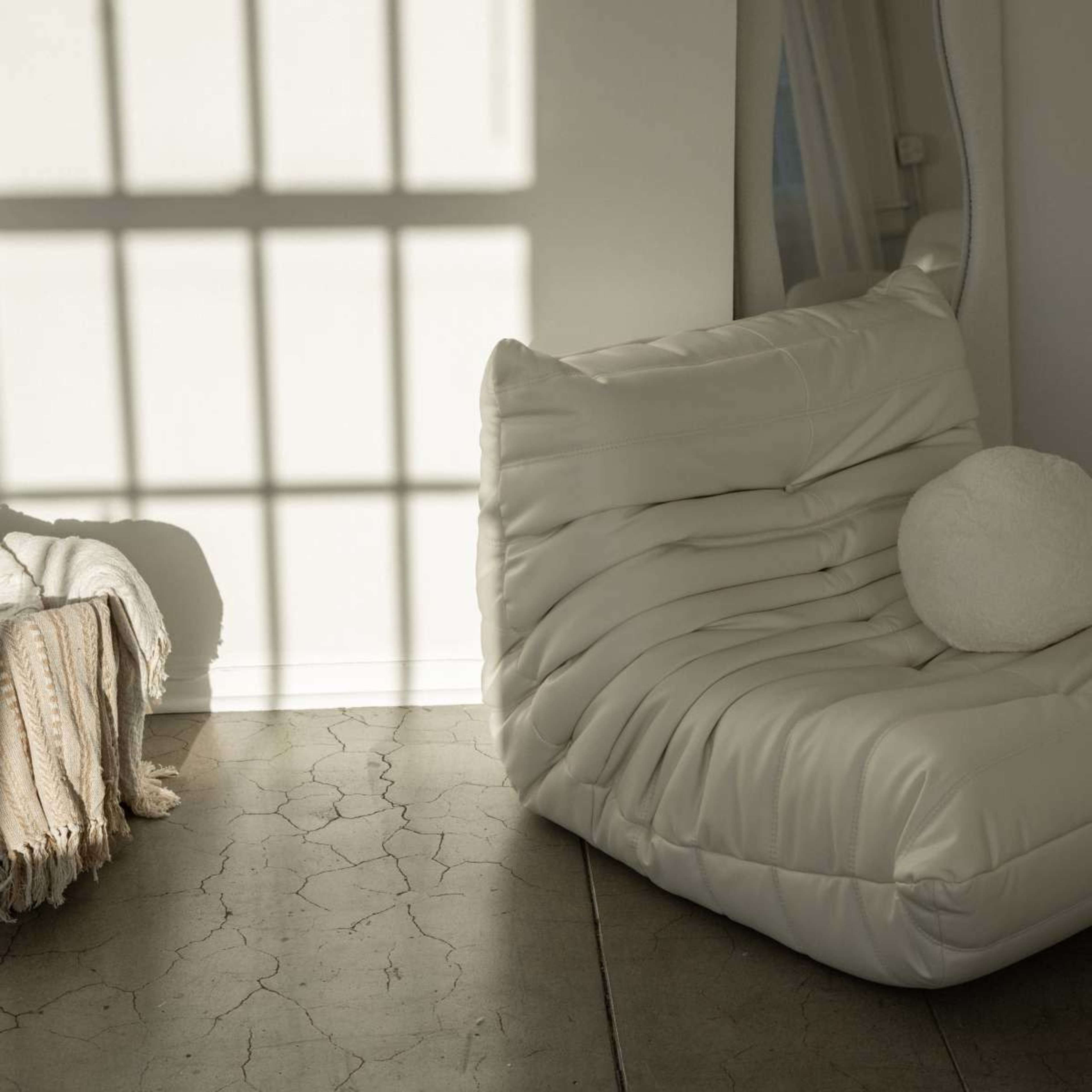 A white, textured lounge chair is positioned beside a draped blanket and casts a shadow from a window onto a cracked floor.