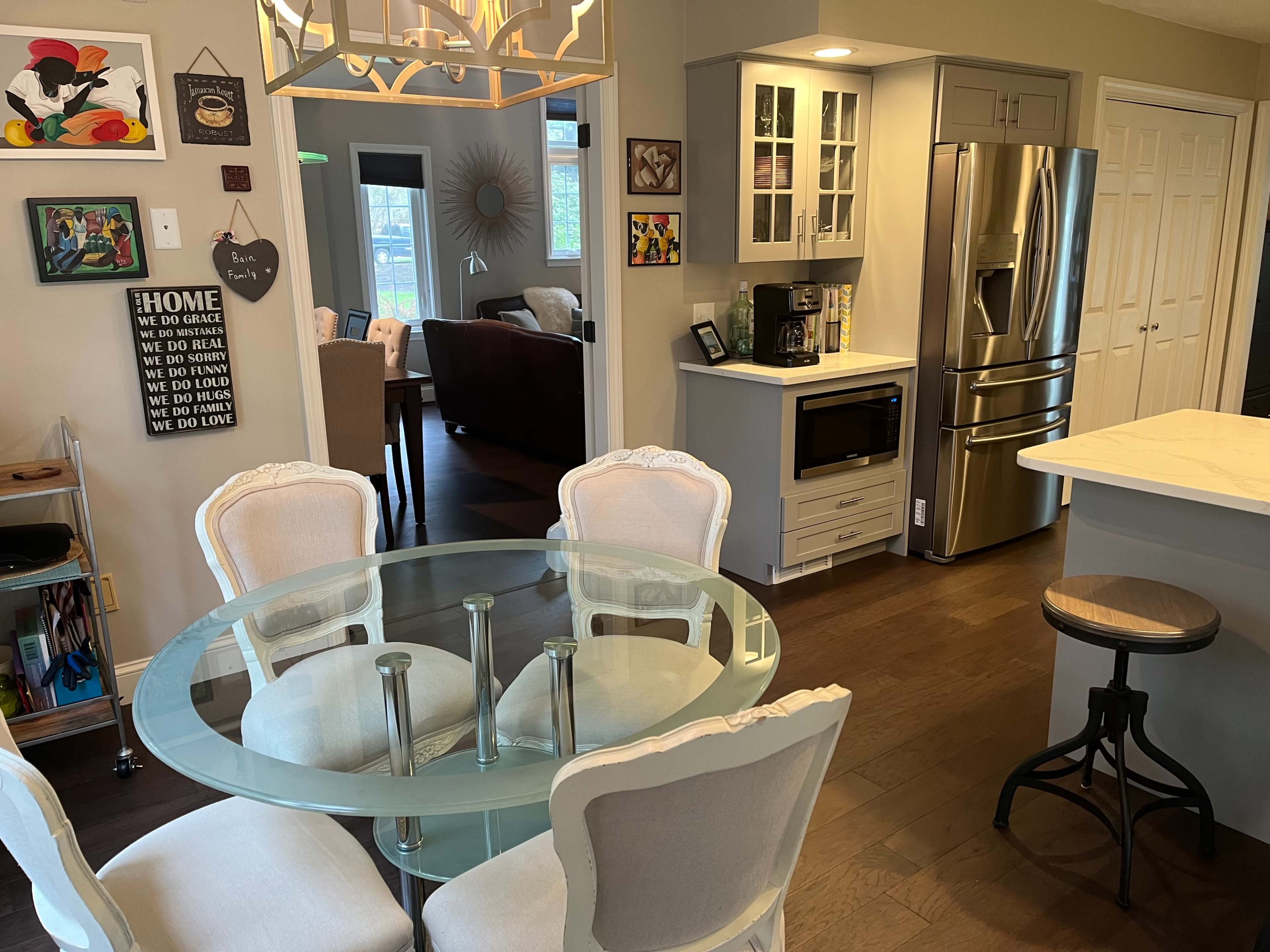 The image shows a modern kitchen area with a glass dining table surrounded by upholstered chairs, alongside a stainless steel refrigerator and a countertop with a microwave.