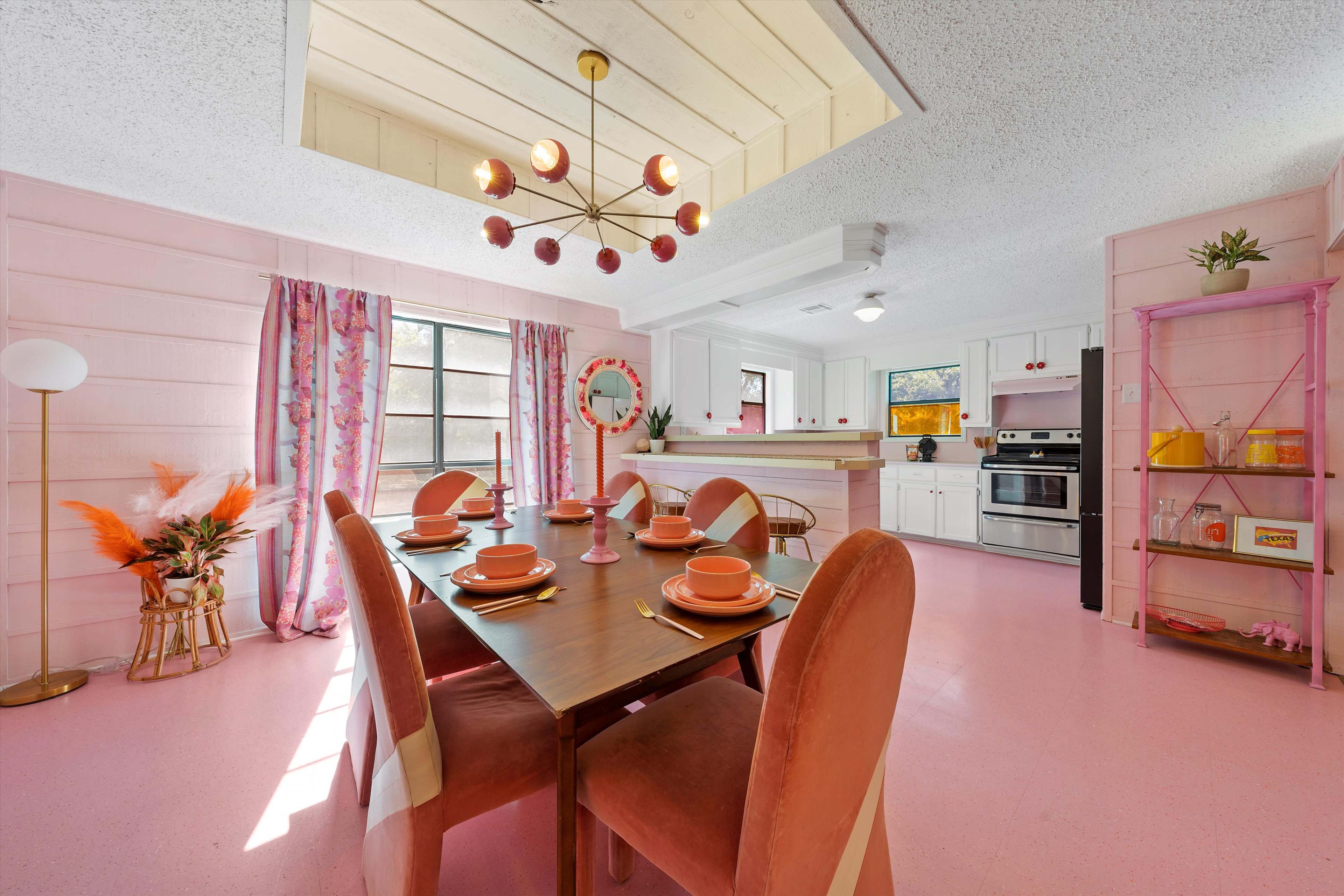 A brightly colored kitchen and dining area with a pink floor, a table set for dinner, and decorative elements like curtains and a chandelier.