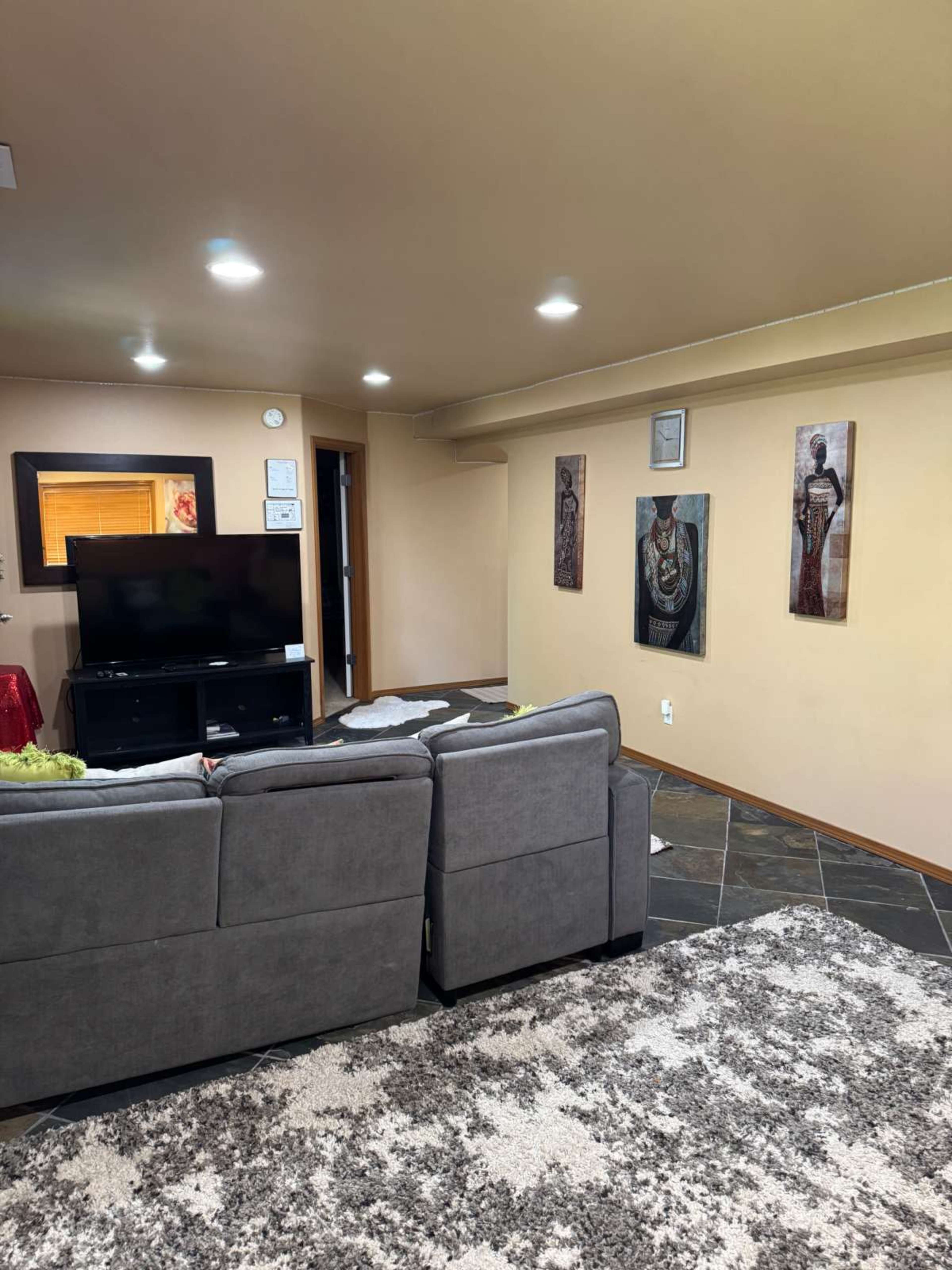 The image shows a living room with a television on a stand, gray sofas facing a wall adorned with framed art, and a rug on a tiled floor.
