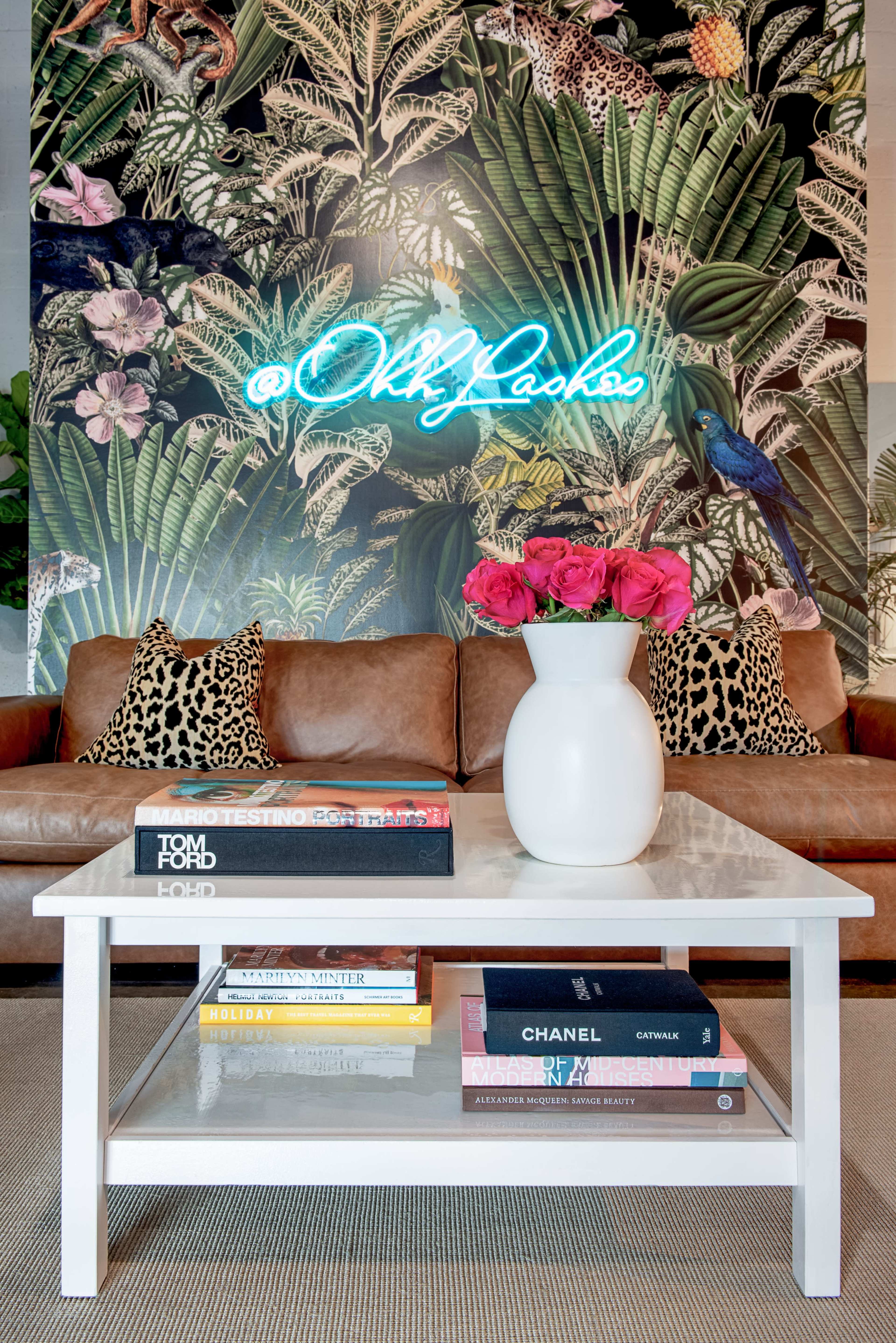 A living room with a brown leather sofa adorned with animal print cushions, a white coffee table stacked with books, and a neon sign against a vibrant, tropical-themed mural.