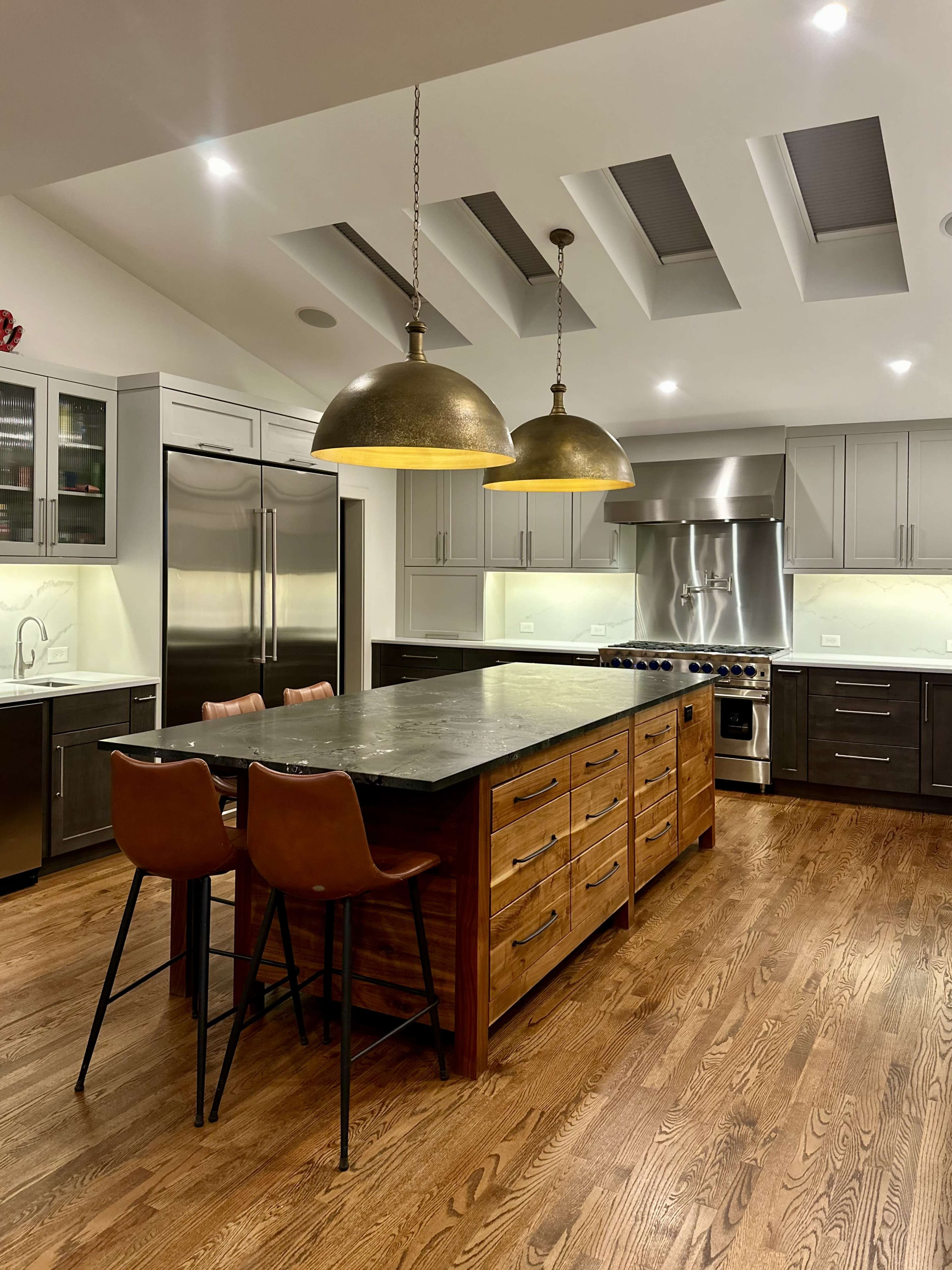 A spacious kitchen features a large central island with wooden drawers, two bar stools, and pendant lighting above, surrounded by modern stainless steel appliances and cabinetry.