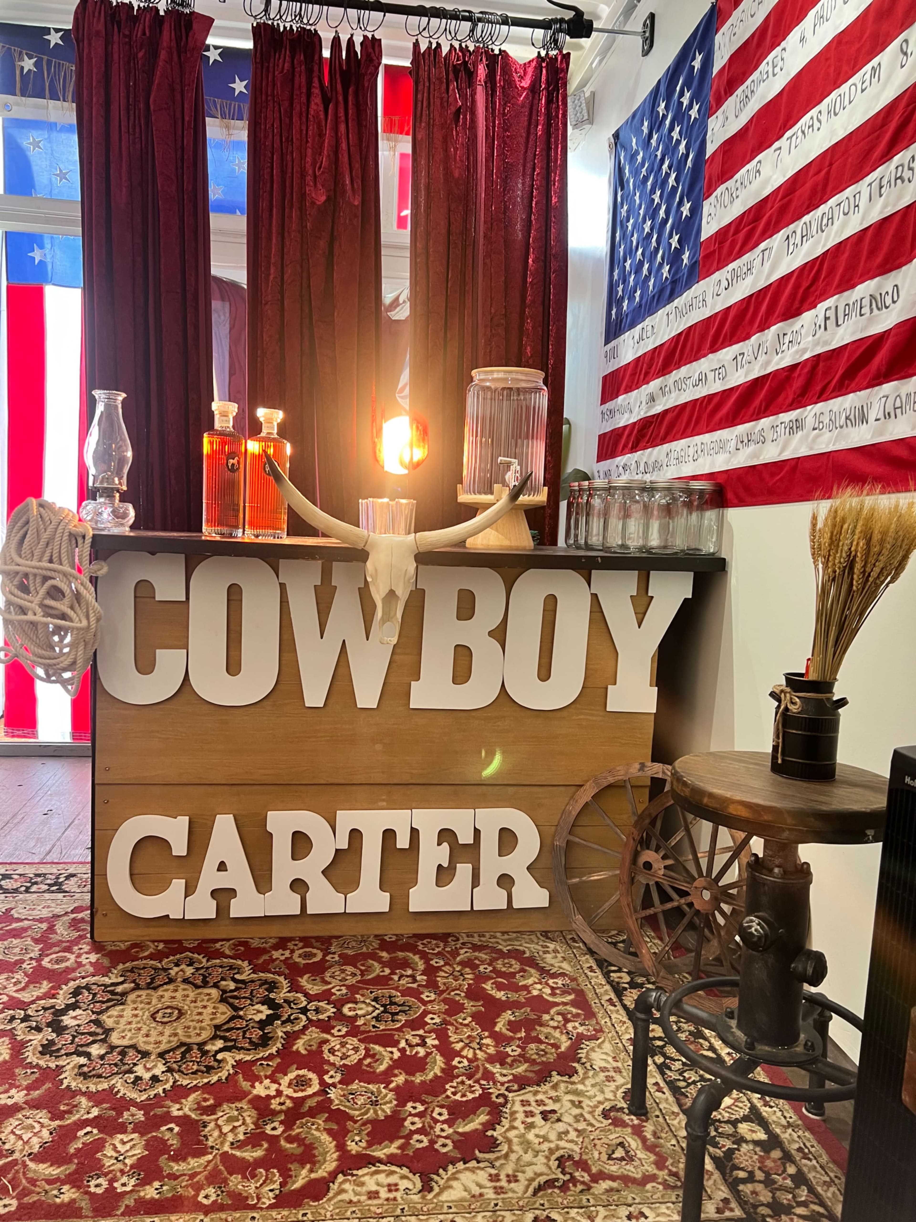 The image shows a rustic bar counter with the words "COWBOY CARTER" prominently displayed in large letters, set against a backdrop of red curtains and an American flag.