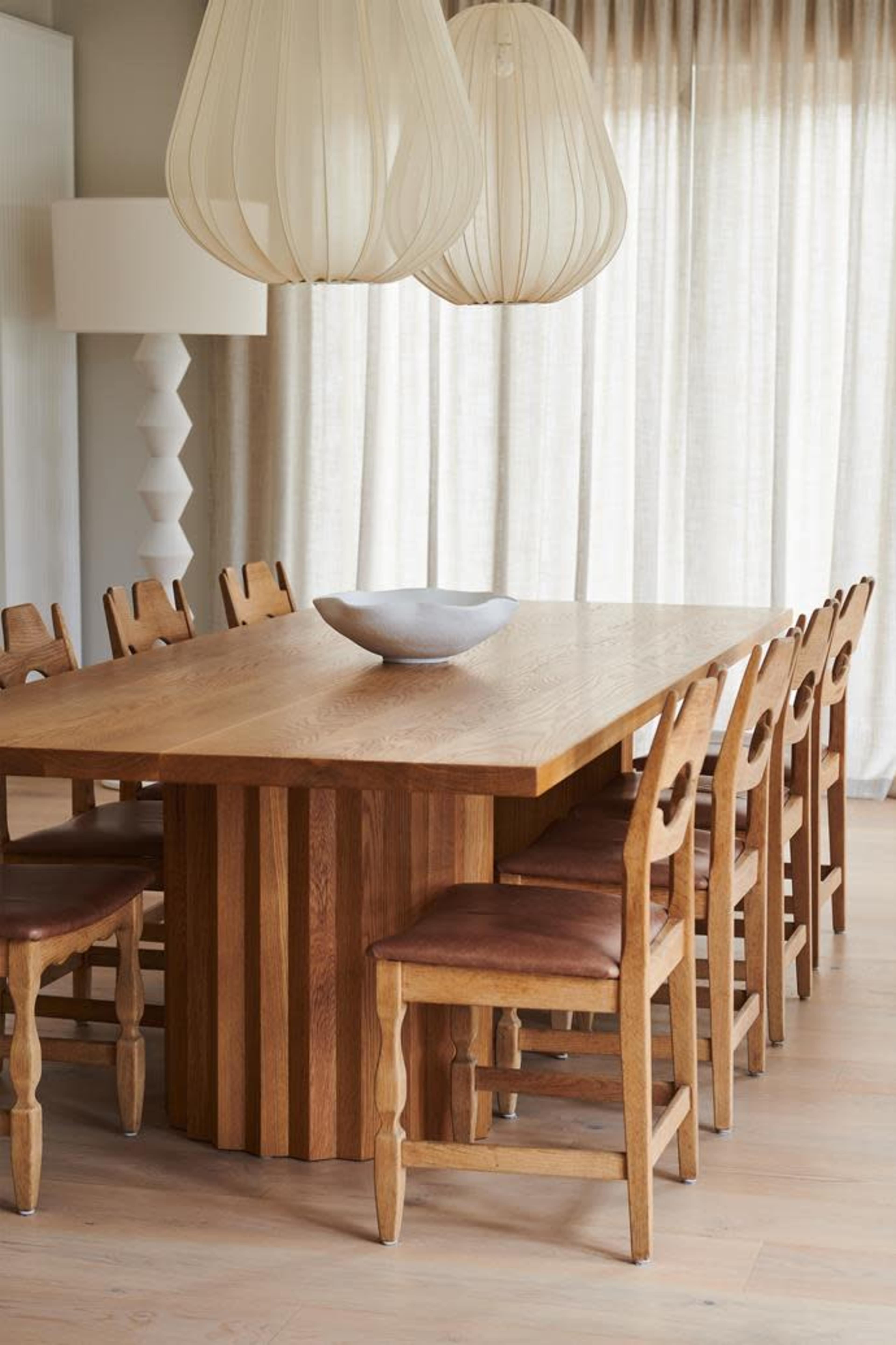 A wooden dining table with a striped pedestal is surrounded by eight upholstered chairs beneath three pendant lights.