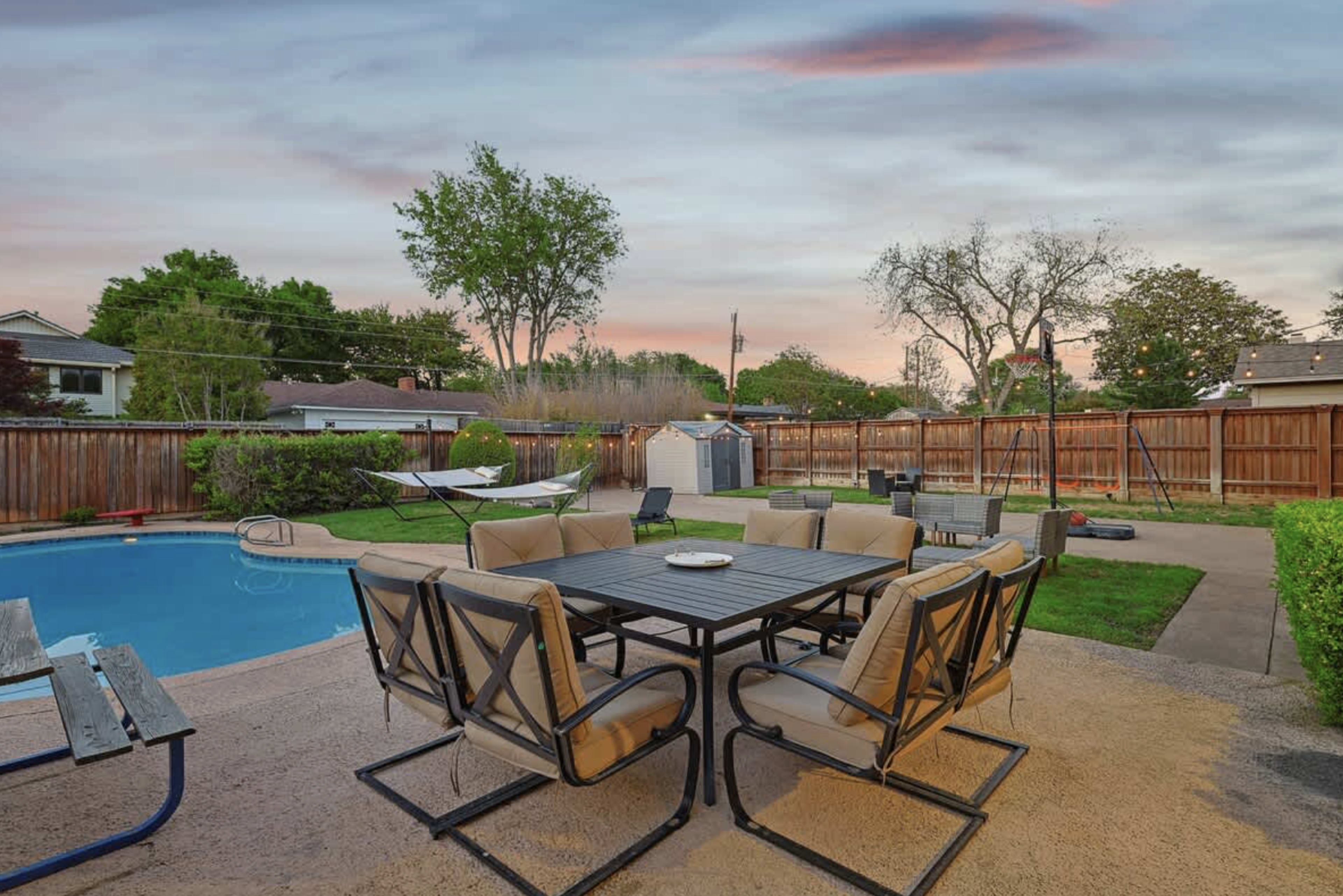 A backyard features a swimming pool, a metal dining set with cushioned chairs, and a hammock surrounded by wooden fencing and trees beneath a sunset sky.