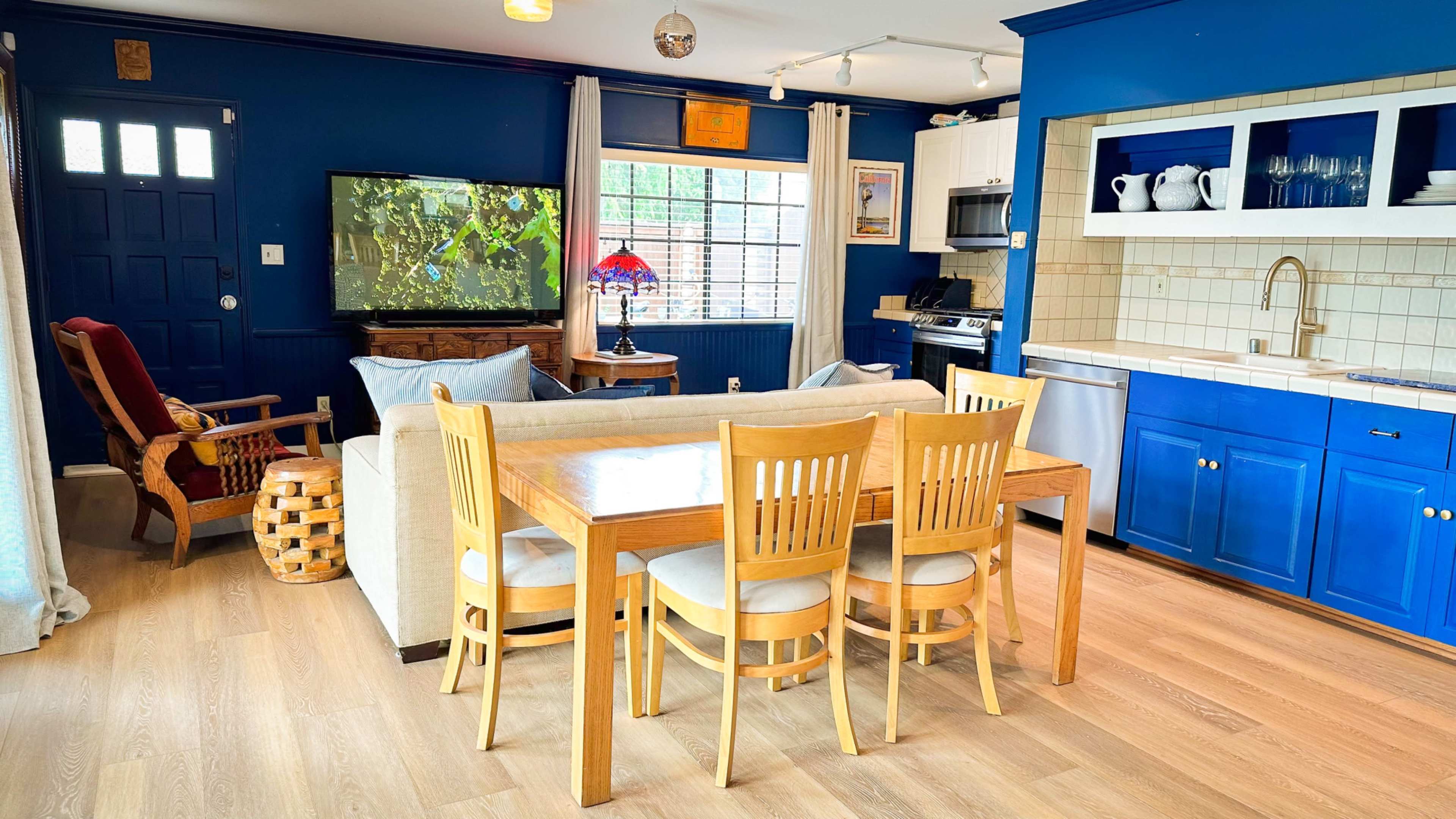 The image shows a bright, modern living space featuring a blue accent wall, a sofa, a dining table with four chairs, and a television mounted on the wall.