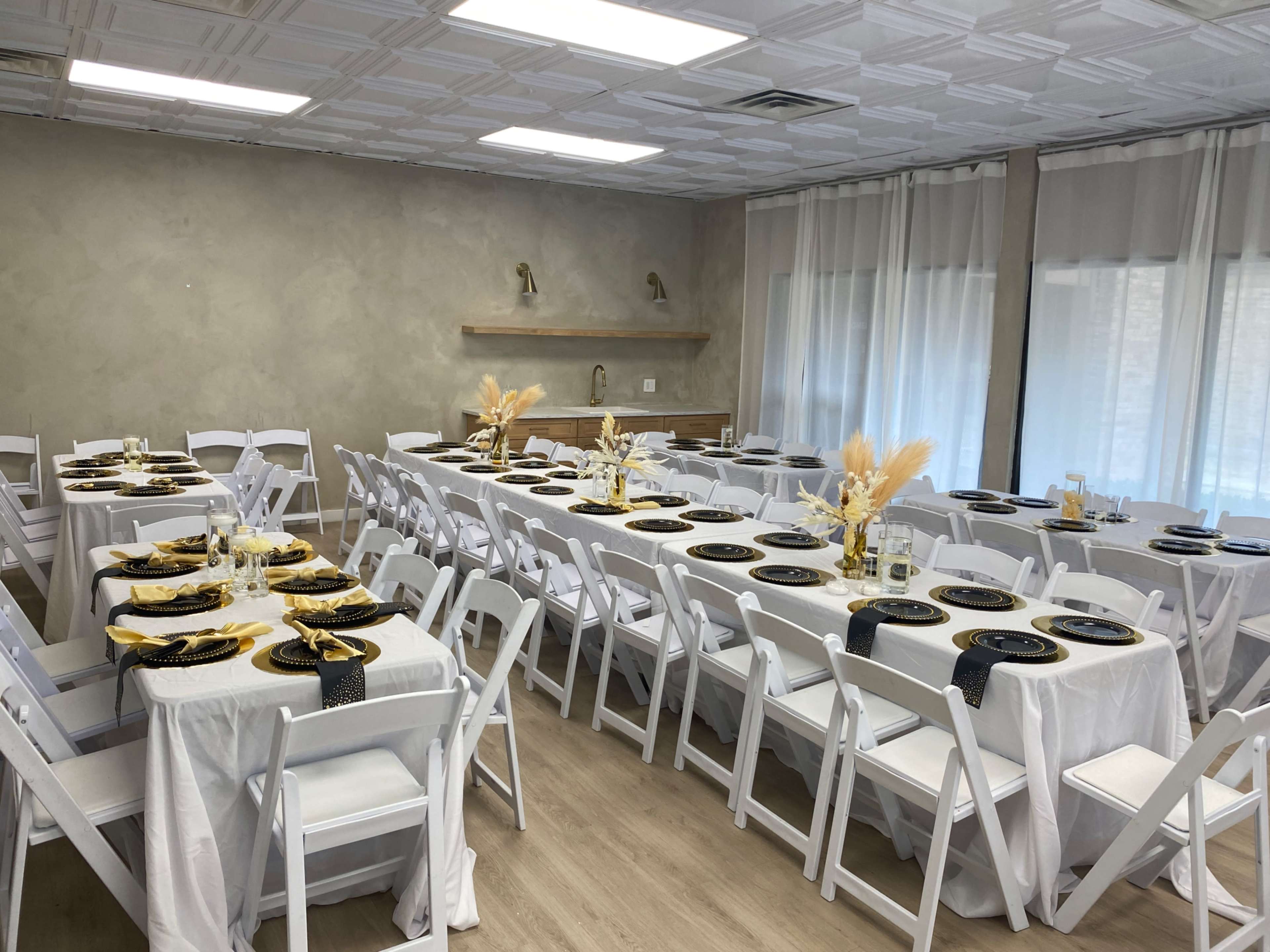 The image shows a banquet hall set up with multiple rectangular tables covered in white cloth, adorned with black plates and decorative centerpieces, arranged neatly to accommodate guests.