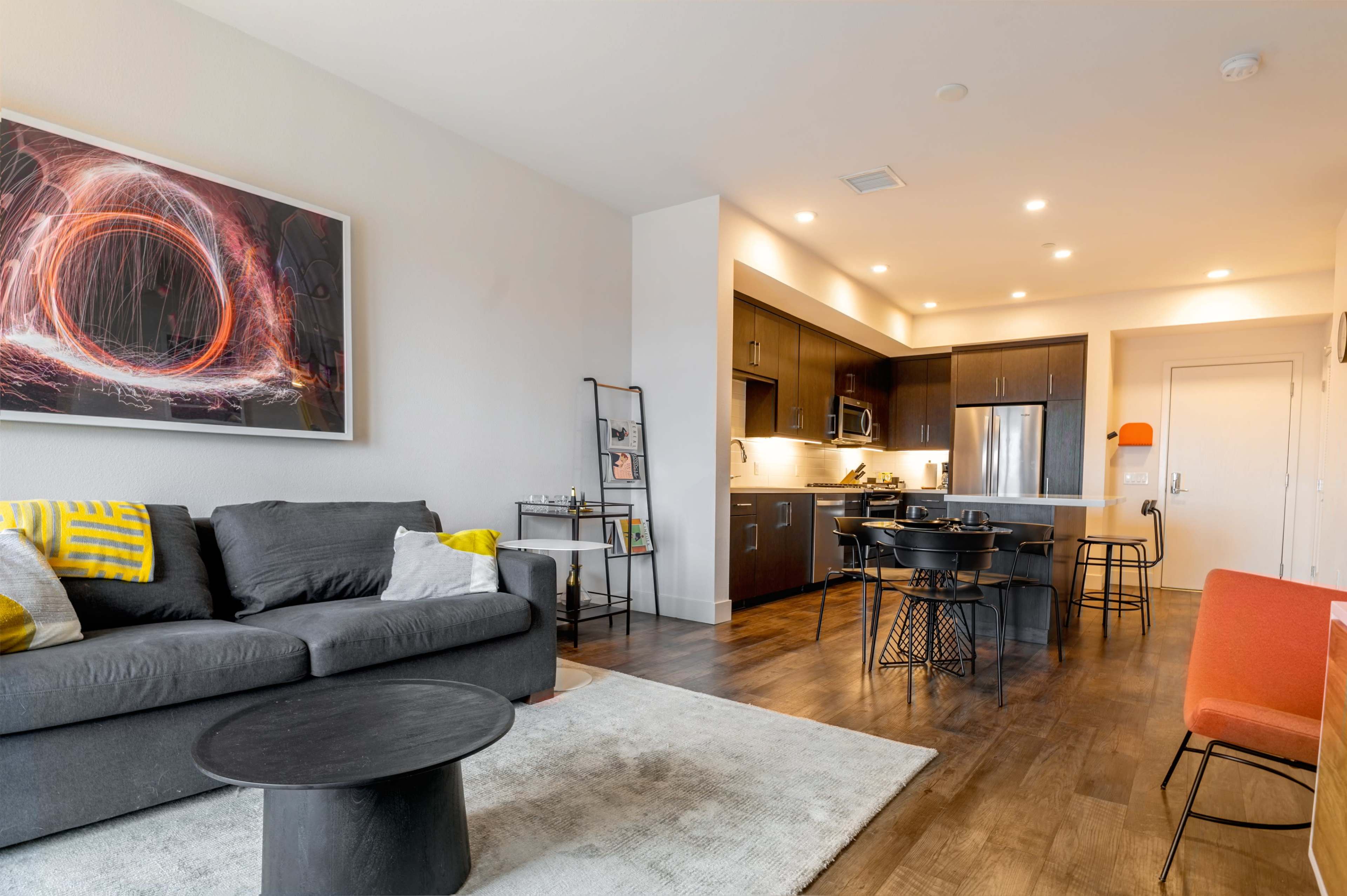 A modern living room with a gray sofa, a round black coffee table, and a kitchen area in the background, featuring dark cabinets and stainless steel appliances.