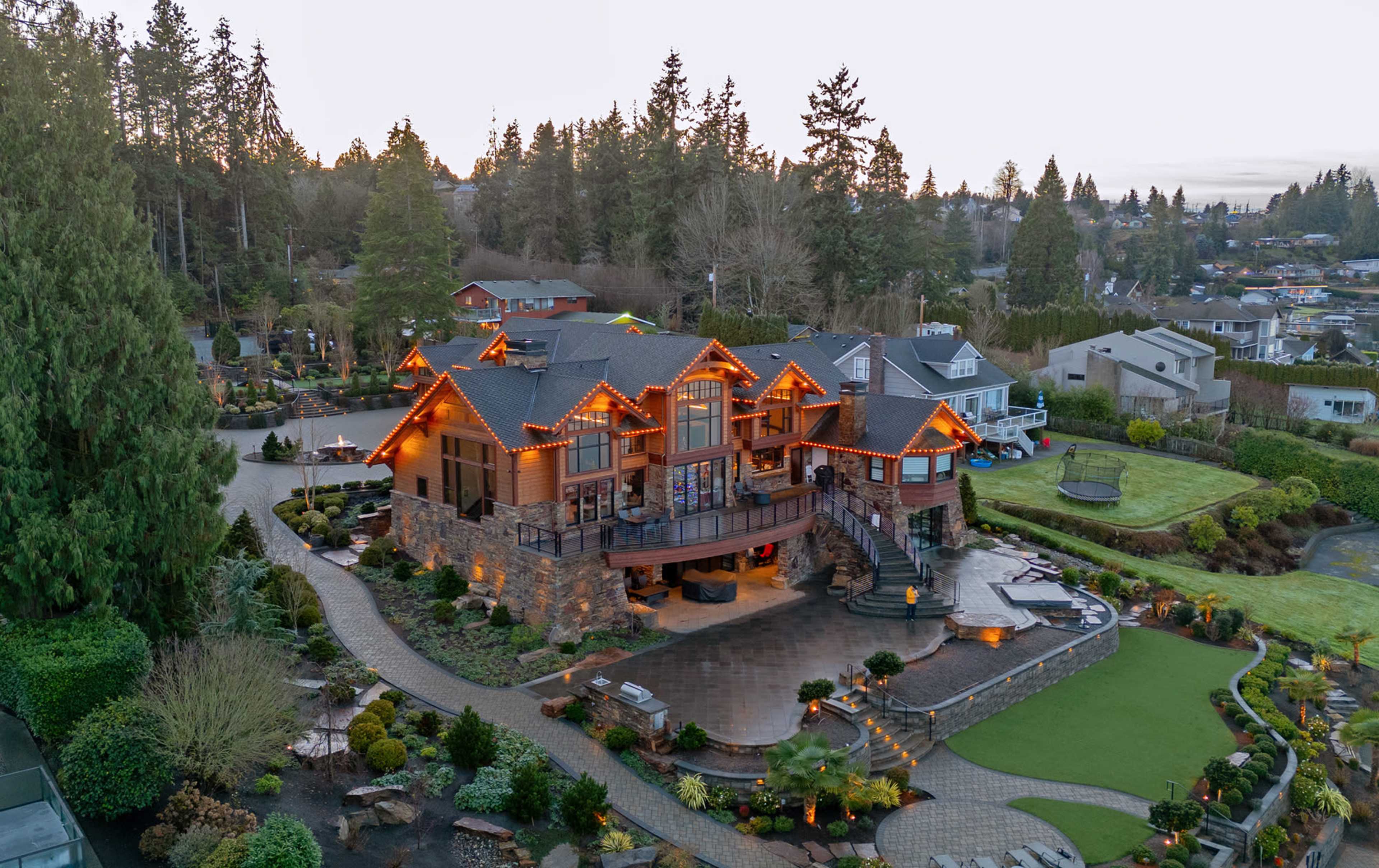 A large, multi-level house decorated with orange lights is surrounded by well-maintained gardens and trees.