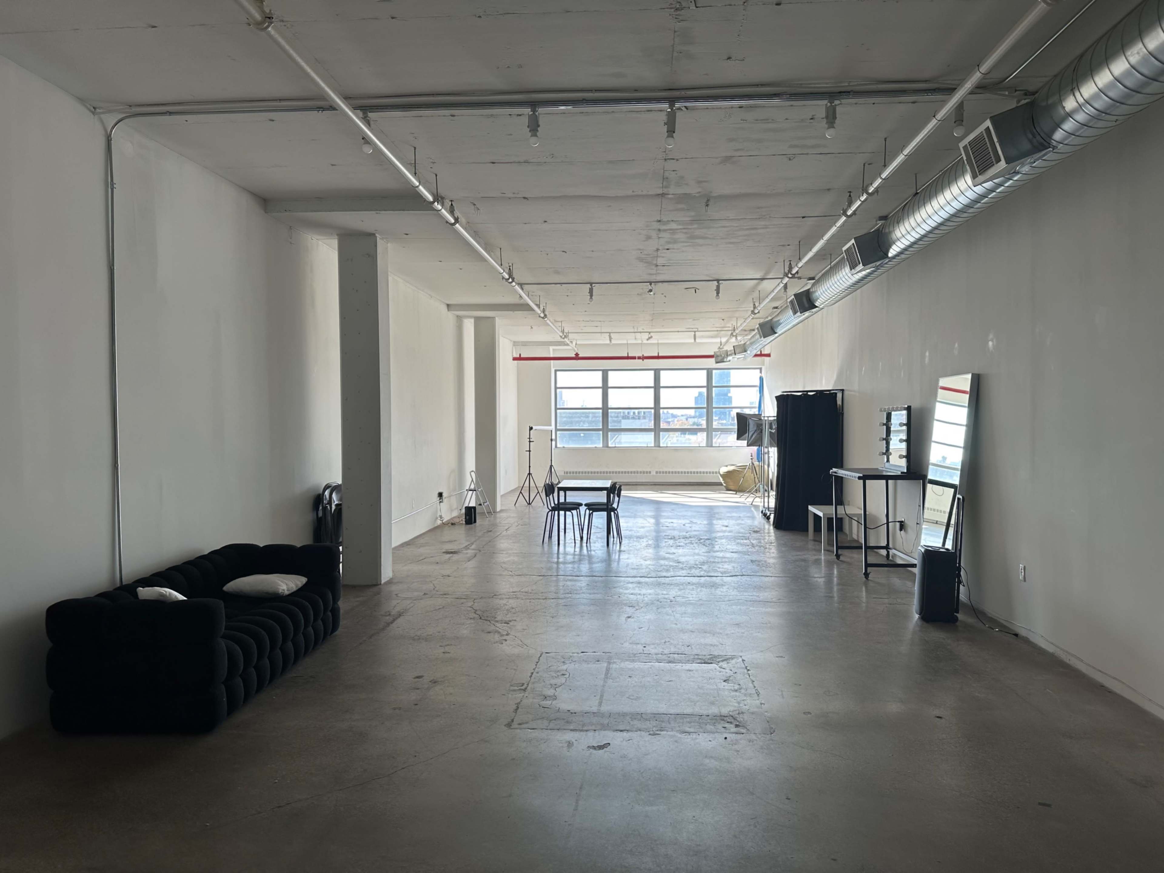 The image shows a spacious, empty room with concrete flooring, large windows allowing natural light, and minimal furnishings including a sofa, a table, and a mirrored cabinet.