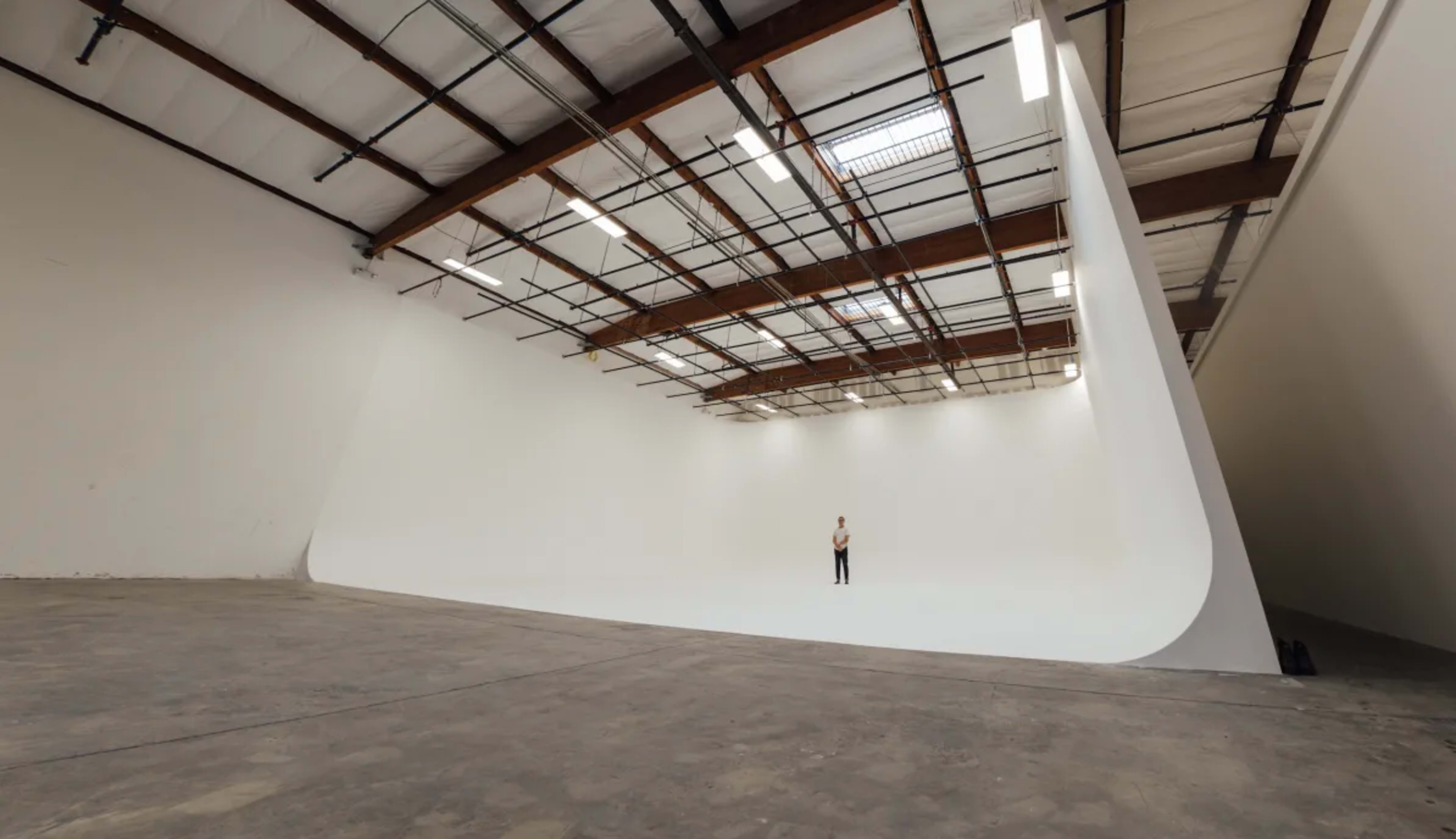 A person stands alone in a large, empty studio with a smooth, curved white backdrop and exposed wooden beams overhead.